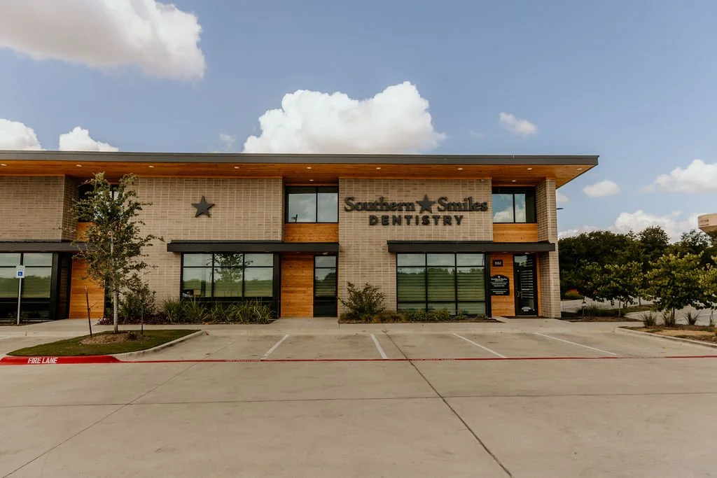 Exterior view of Southern Smiles Dentistry building with a parking lot in front, trees on either side, and a clear sky with some clouds above.