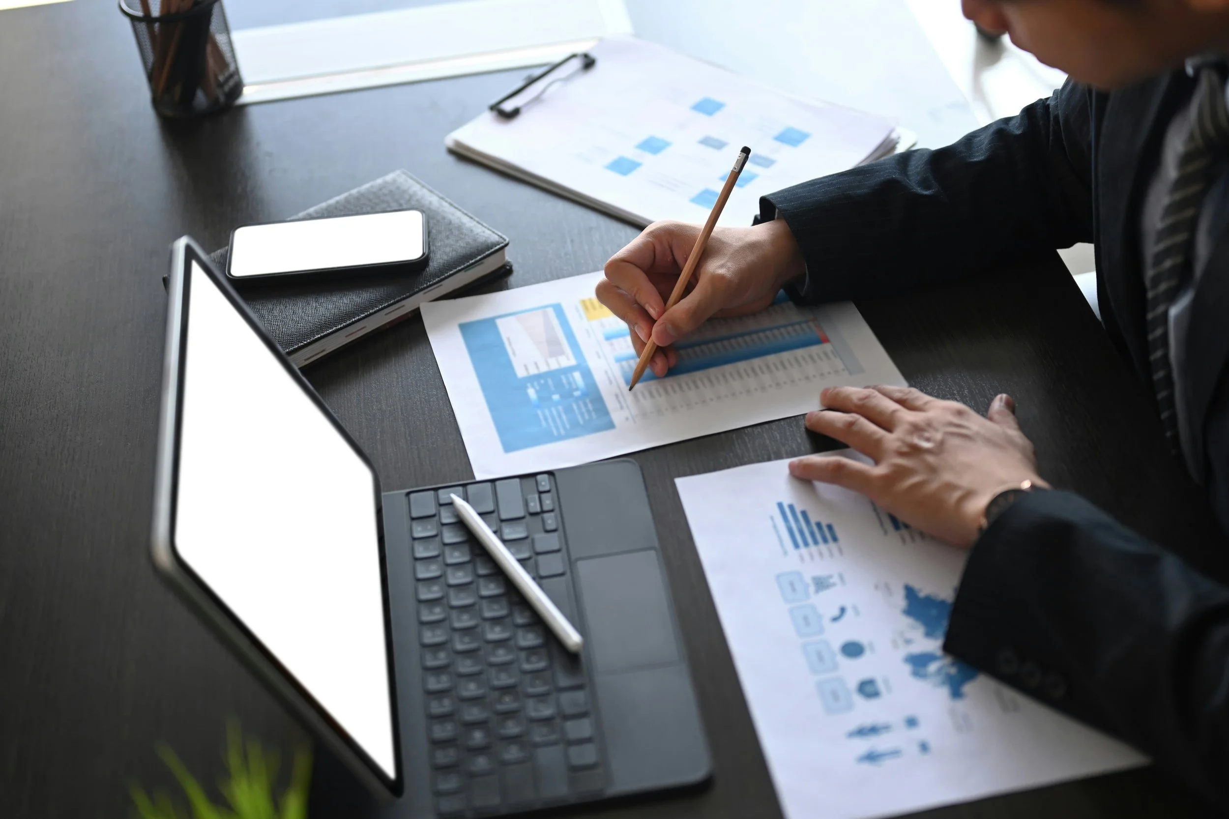 Person in business attire working on printed financial charts and graphs at a desk, with electronic devices and documents around.