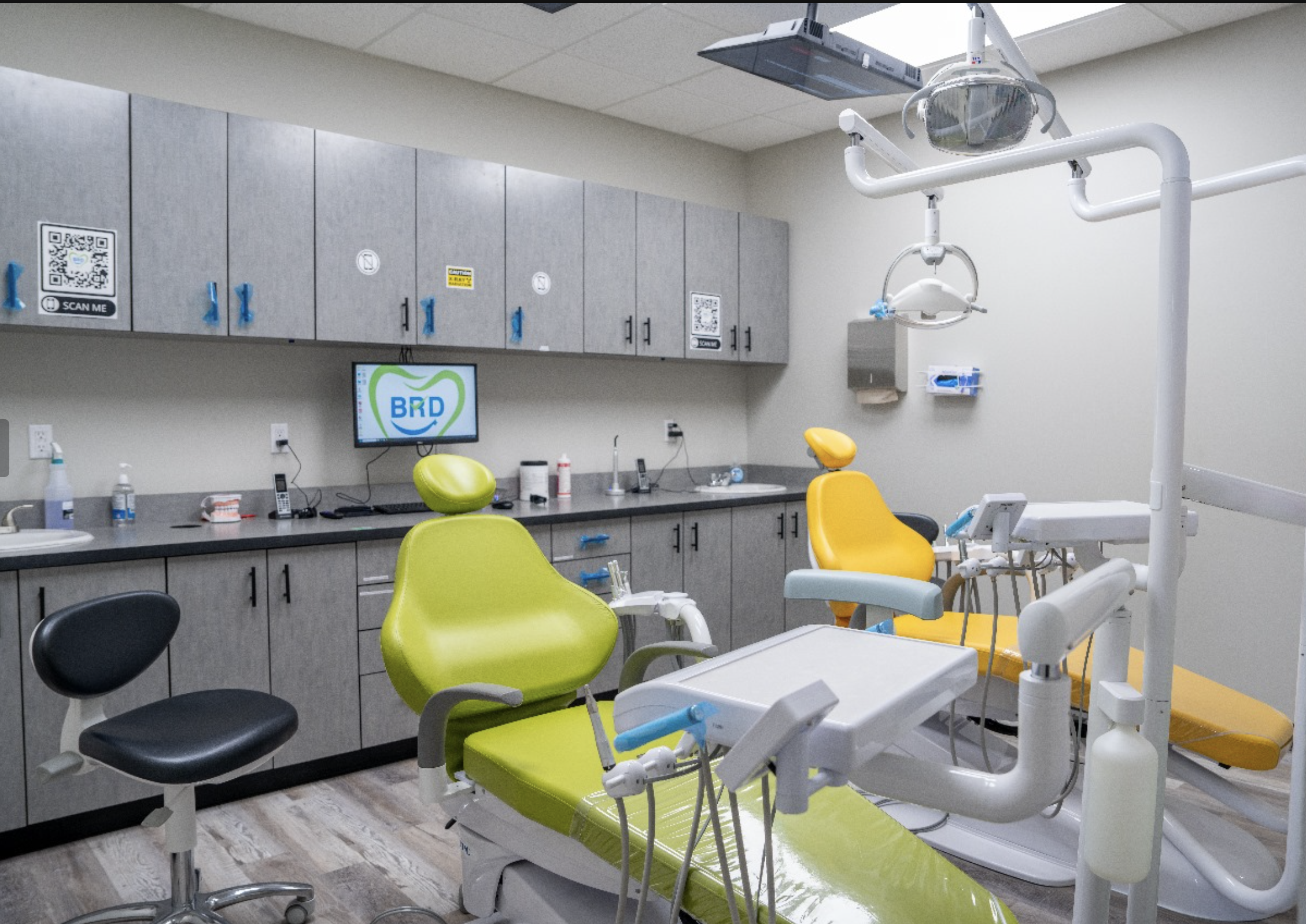 Dental clinic room with two dental chairs, one green and one yellow, equipped with dental tools and overhead lights. Cabinets and a computer monitor displaying 'BRD' are visible in the background.
