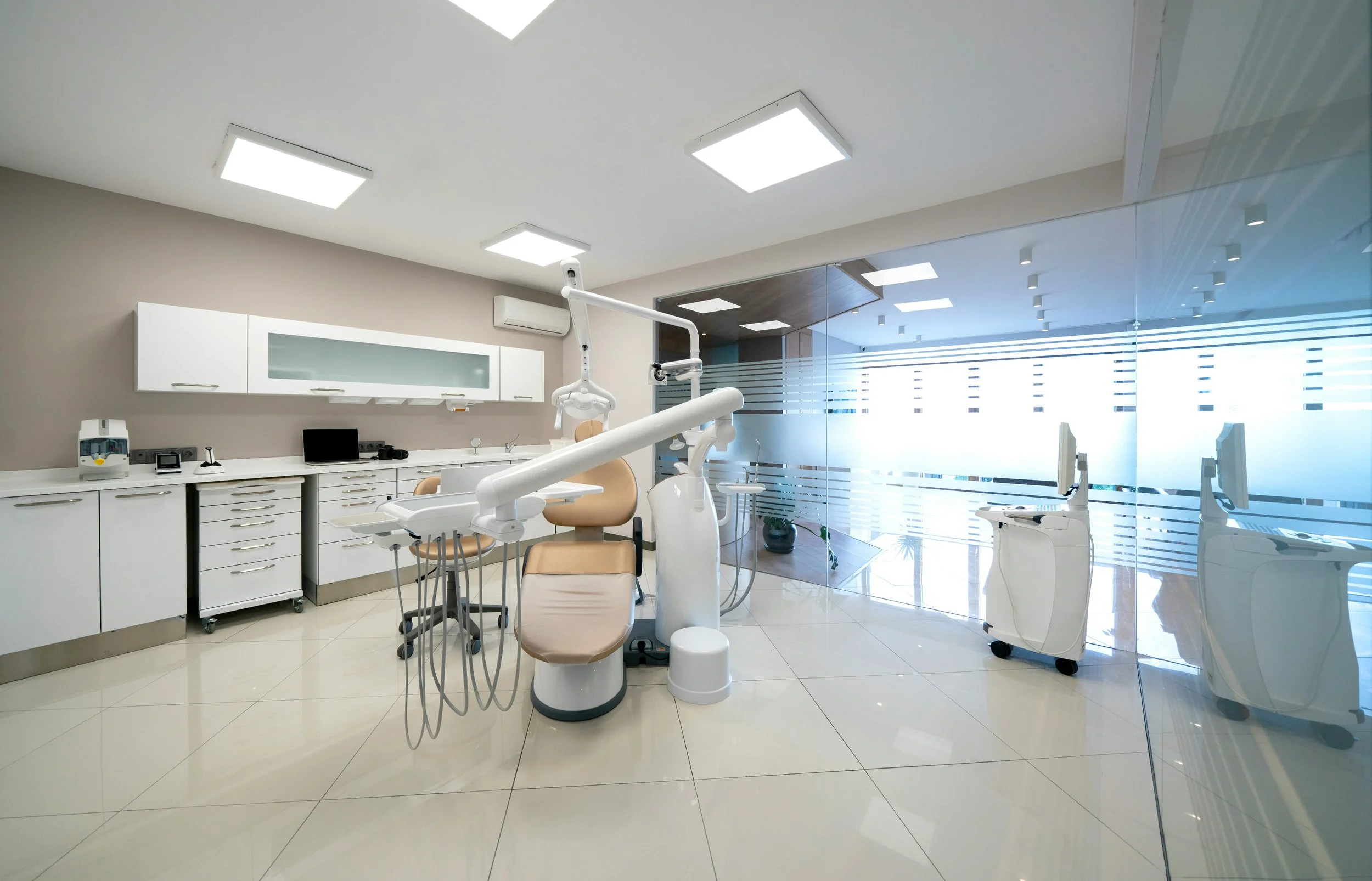 Modern dentist office with dental chair, overhead light, and dental equipment, separated by glass wall with frosted stripes.