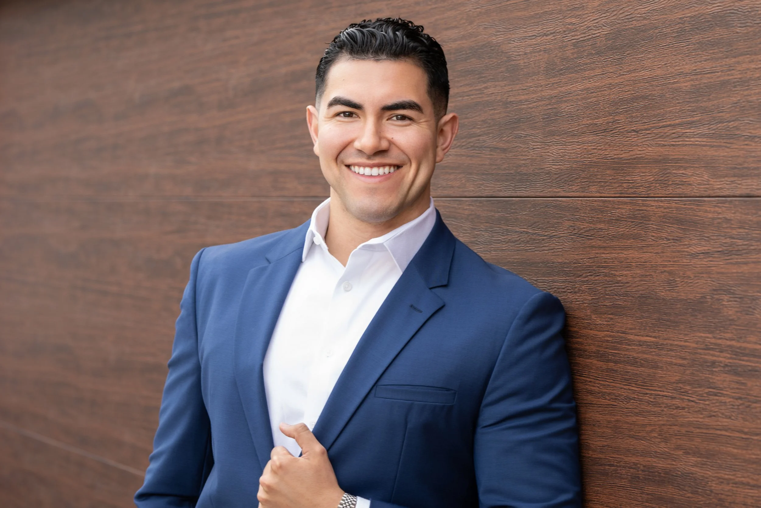 A smiling man in a blue suit with a white shirt, standing against a wooden wall.