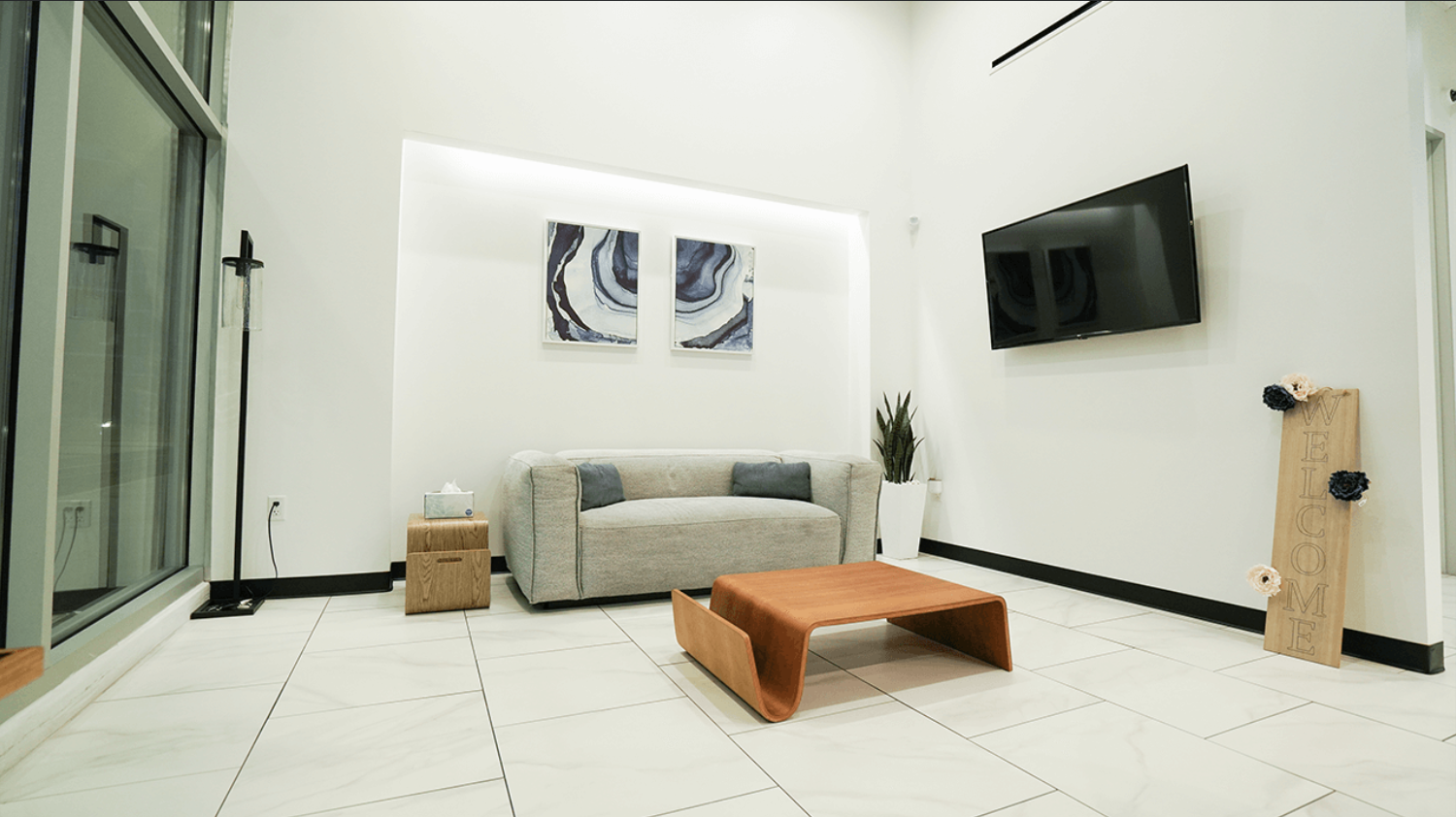 Modern living room with white walls, a gray sofa with two gray pillows, a wooden coffee table, a potted plant, wall art, a wall-mounted TV, and a "Welcome" wooden sign with flowers