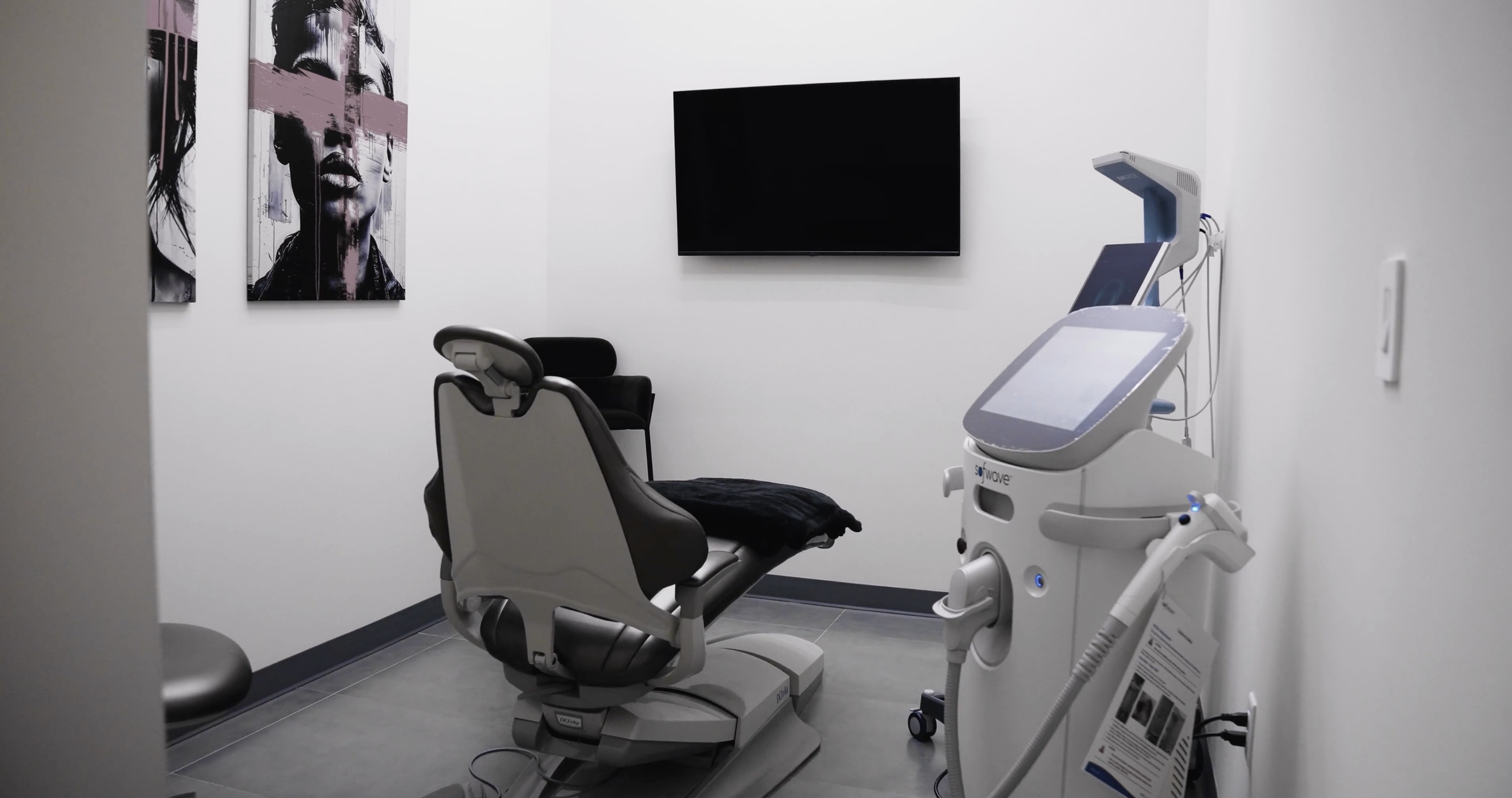 Medical examination room with a gynecological exam chair, ultrasound machine, wall-mounted TV, and artwork of a woman's portrait on the walls.