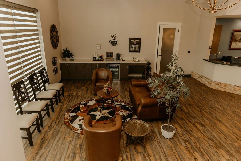 A cozy waiting room with a leather sofa, chairs, a round coffee table, a round patterned rug, and a potted plant near the sofa. There's a window with horizontal blinds, wall decorations, and a countertop with a coffee station.