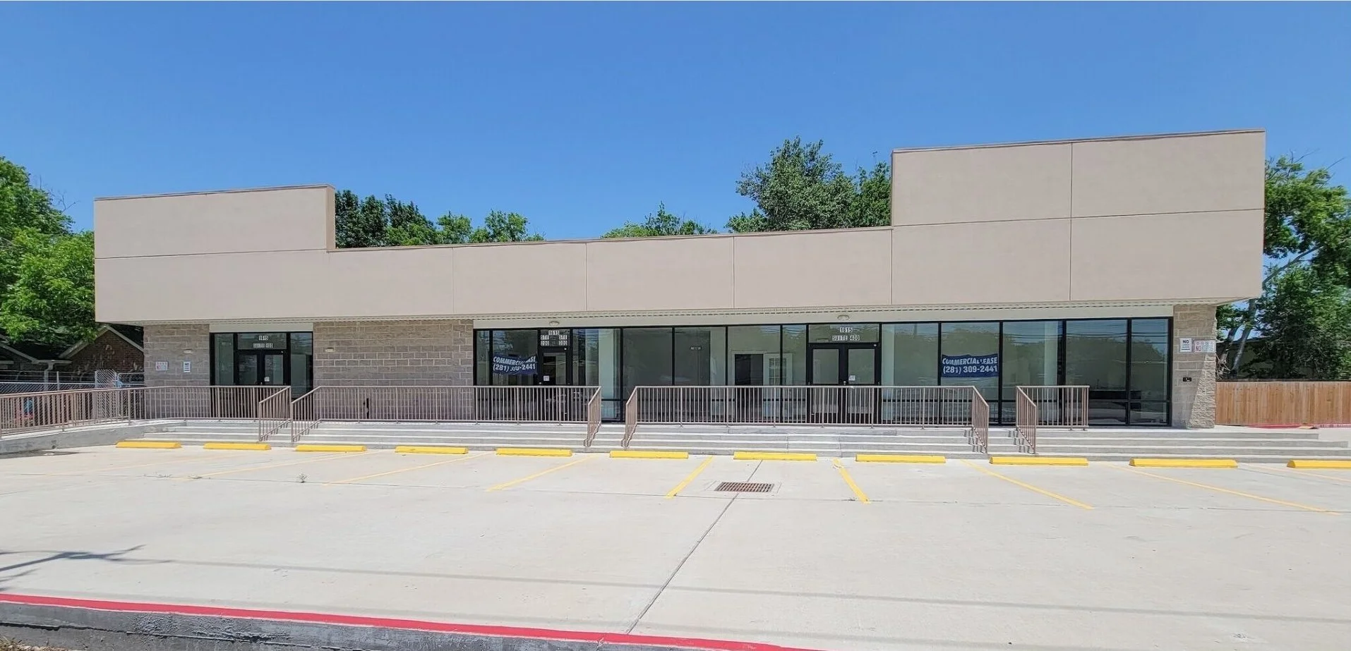 Empty commercial building with a glass storefront and a paved parking lot in front, under a blue sky with green trees in the background.