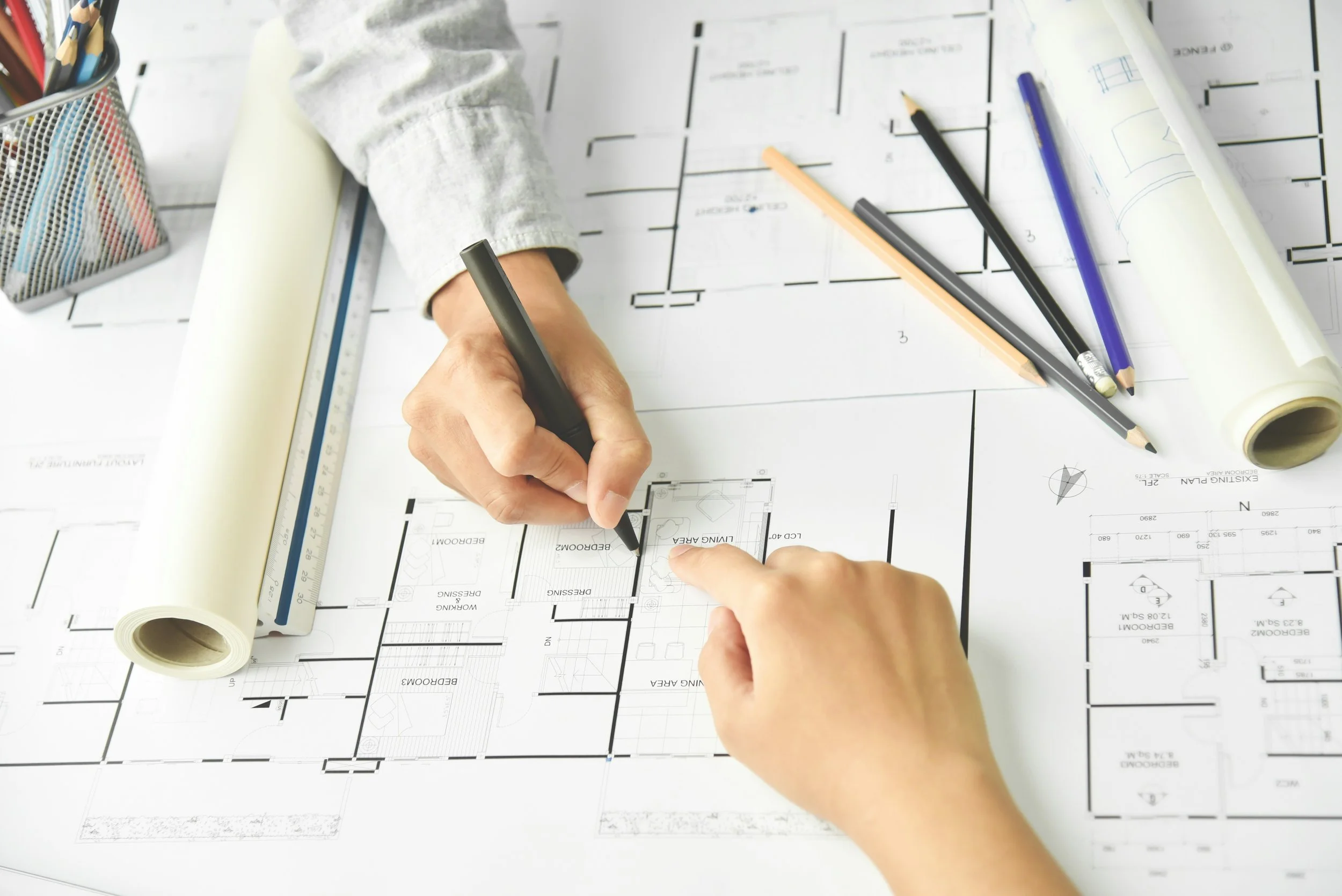 Architect or designer pointing at a blueprint layout on a desk, with various pencils, pens, a ruler, and rolled-up blueprints nearby.
