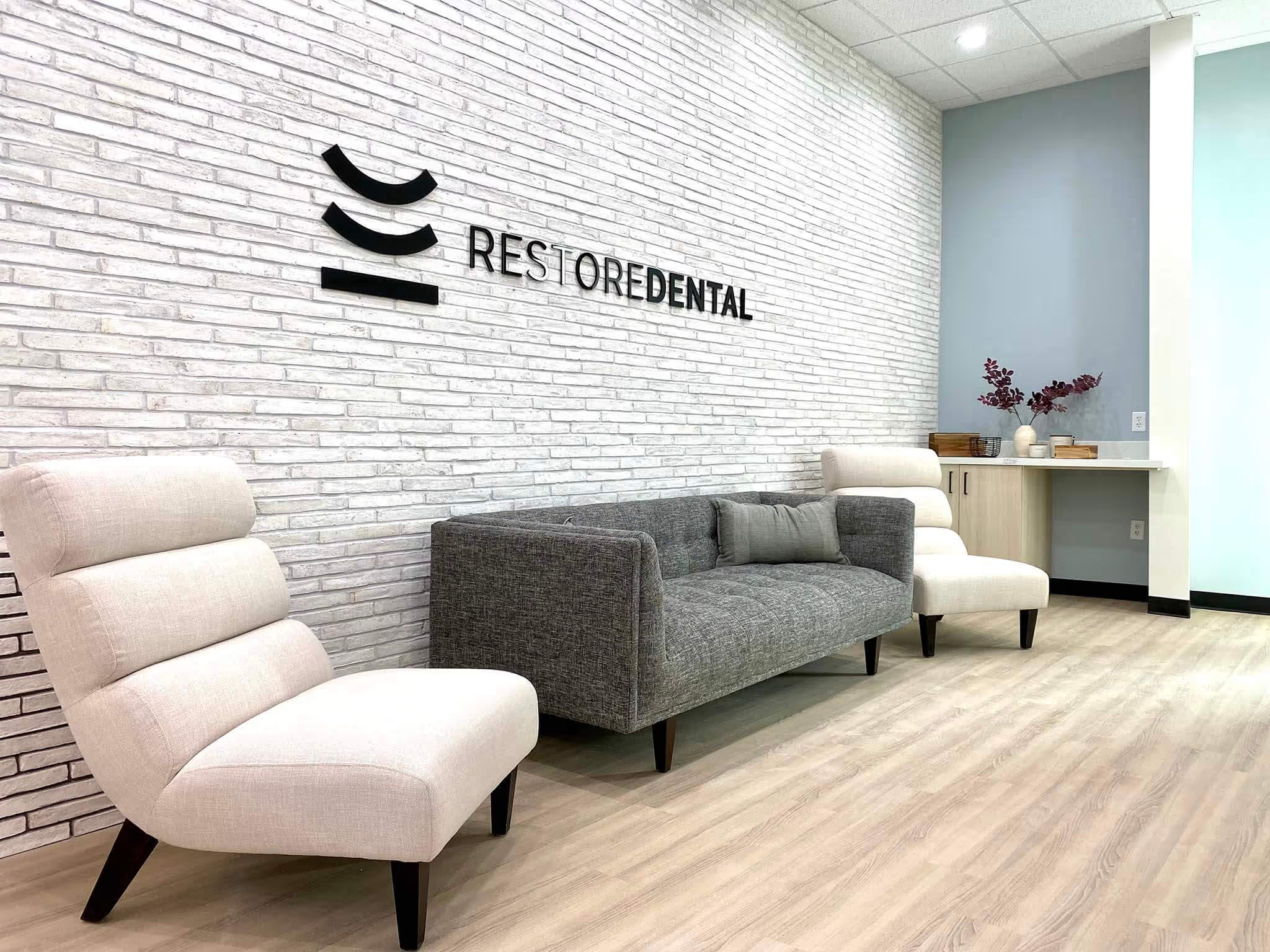 Modern dental office reception area with white brick wall, black signage that says 'Restore Dental', beige and gray chairs, wooden floor, and a countertop with a vase of purple leaves and decorative items.