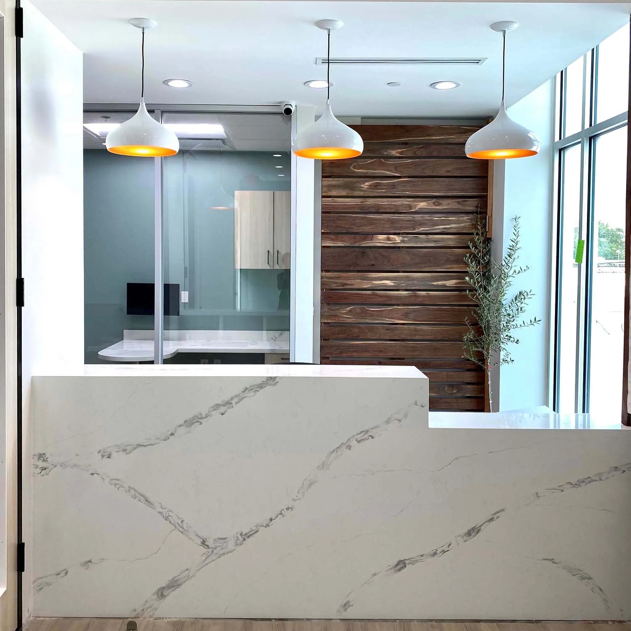Modern reception desk with white marble surface, three white pendant lights hanging from the ceiling, a wooden accent wall, a potted plant, and large glass windows letting in natural light.