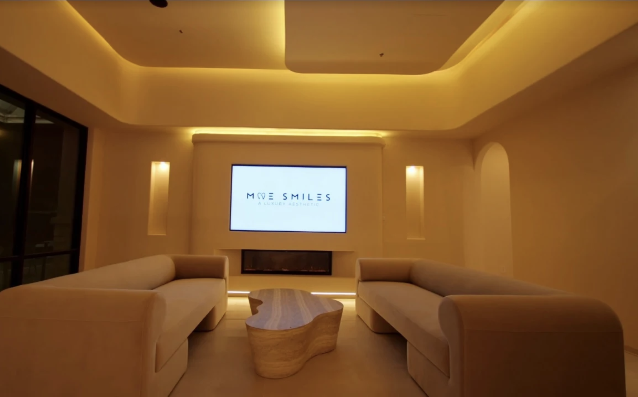 Modern hotel lounge with cream-colored sofas, a wooden coffee table, and a large television screen displaying 'Mole Smiles, A Luxury Aesthetic.' Soft yellow lighting creates an inviting atmosphere.
