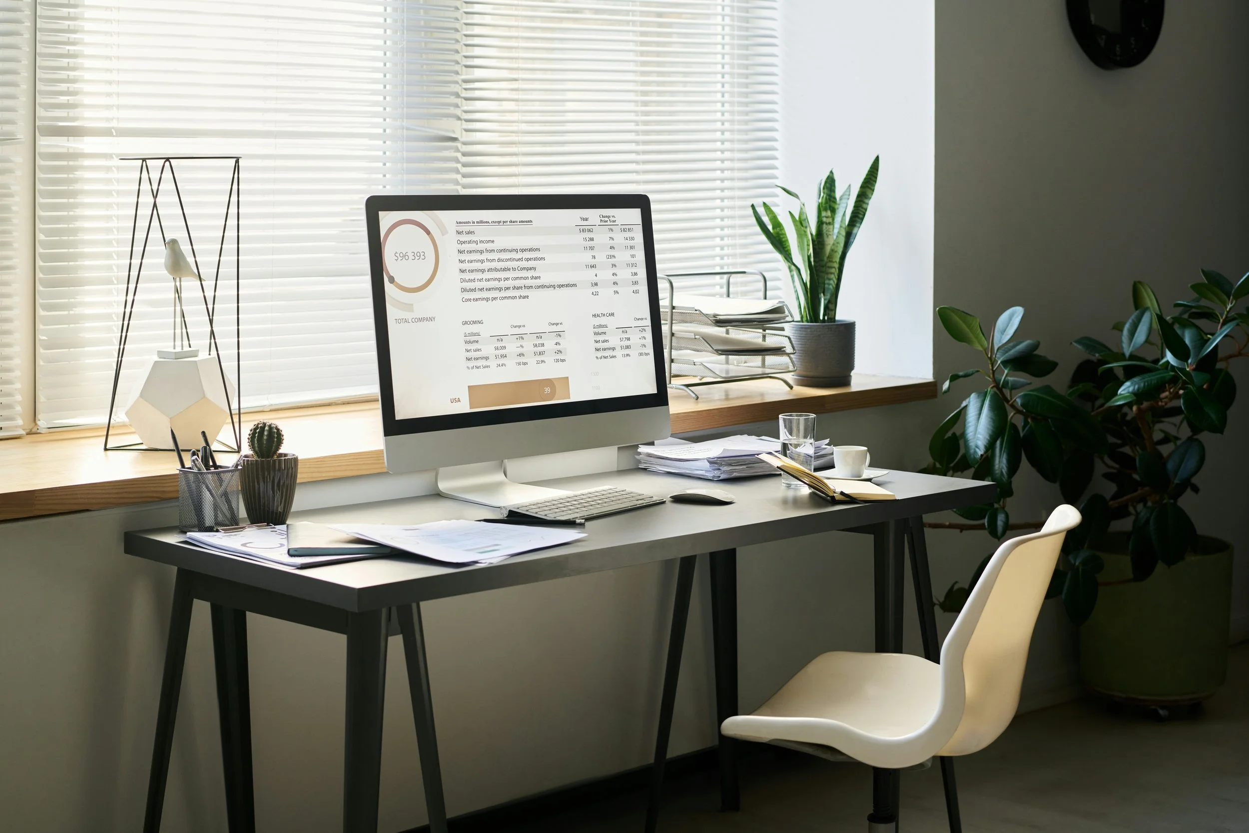 Office desk with a computer monitor, stacks of papers, a glass of water, a cup, and plants near a window with blinds.