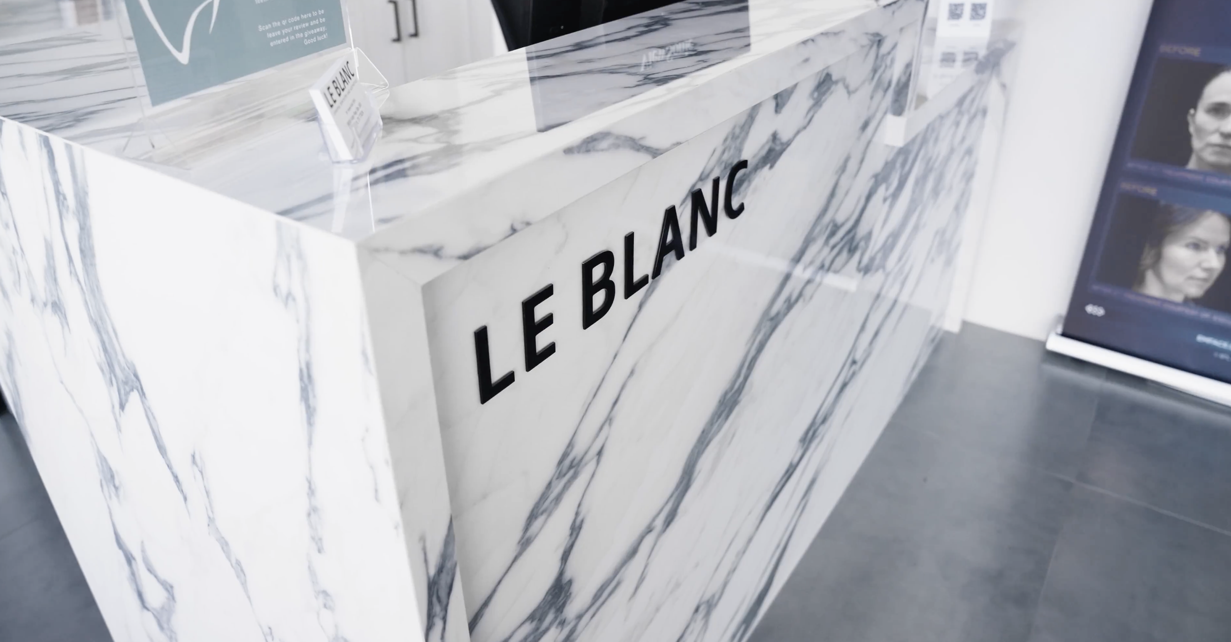 A marble reception desk with the words 'LE BLANC' in black letters, located in a modern, bright space, with a poster in the background and a digital screen displaying face images to the right.