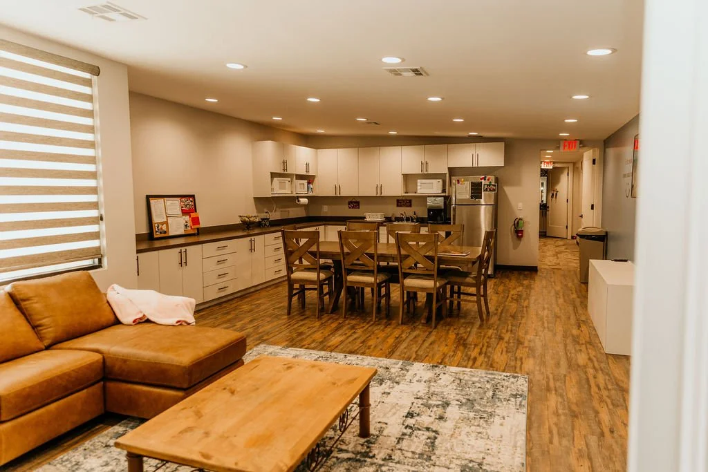 Open kitchen and living room with wooden flooring, beige cabinets, a dining table with eight chairs, a brown leather sofa, a wooden coffee table, and windows with striped blinds.