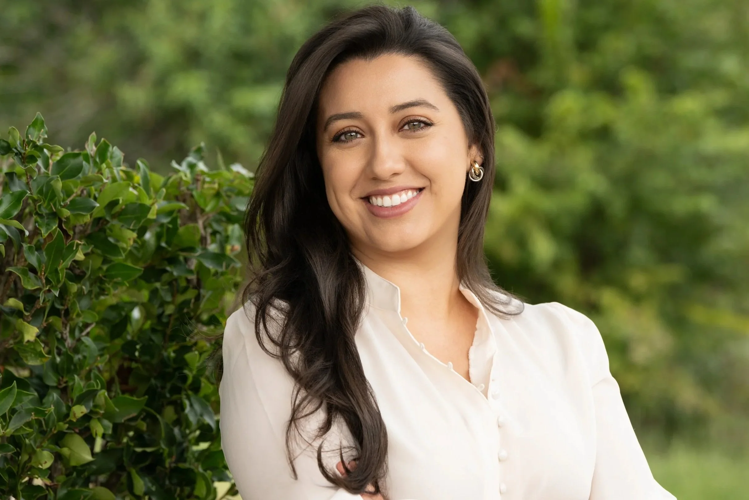 A woman with long dark hair and earrings, smiling in front of green foliage, wearing a white blouse.