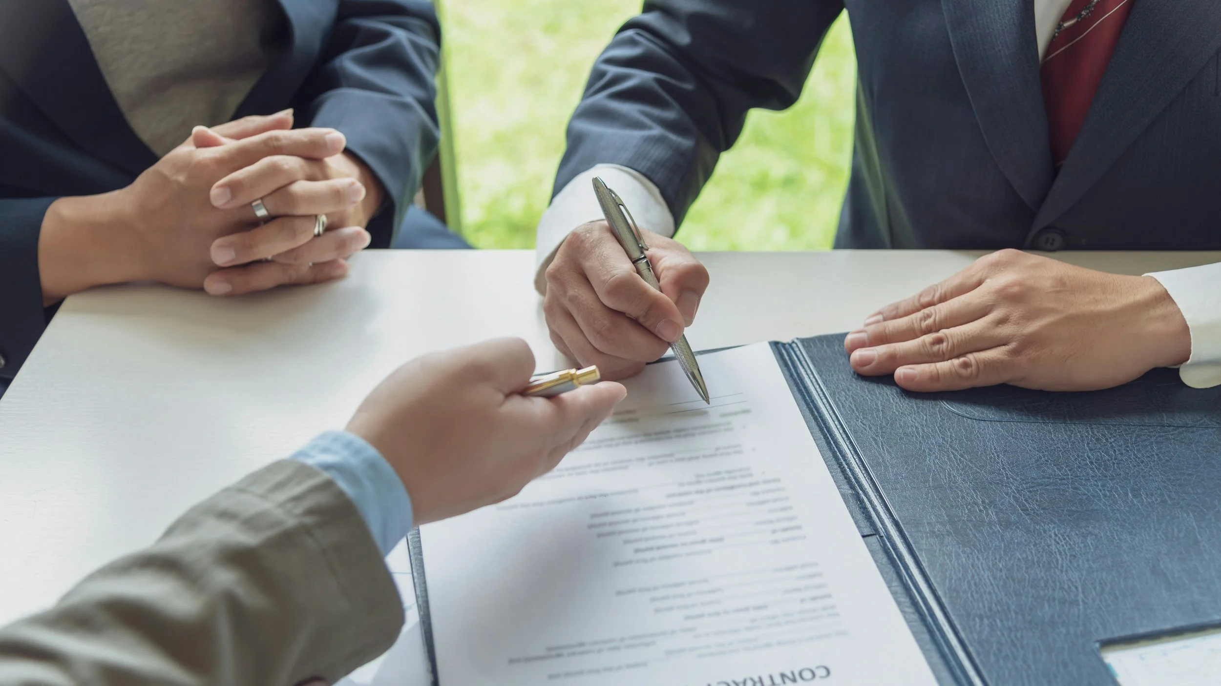 Two people signing a contract document on a desk with one person holding a pen and the other resting their hand on the document.