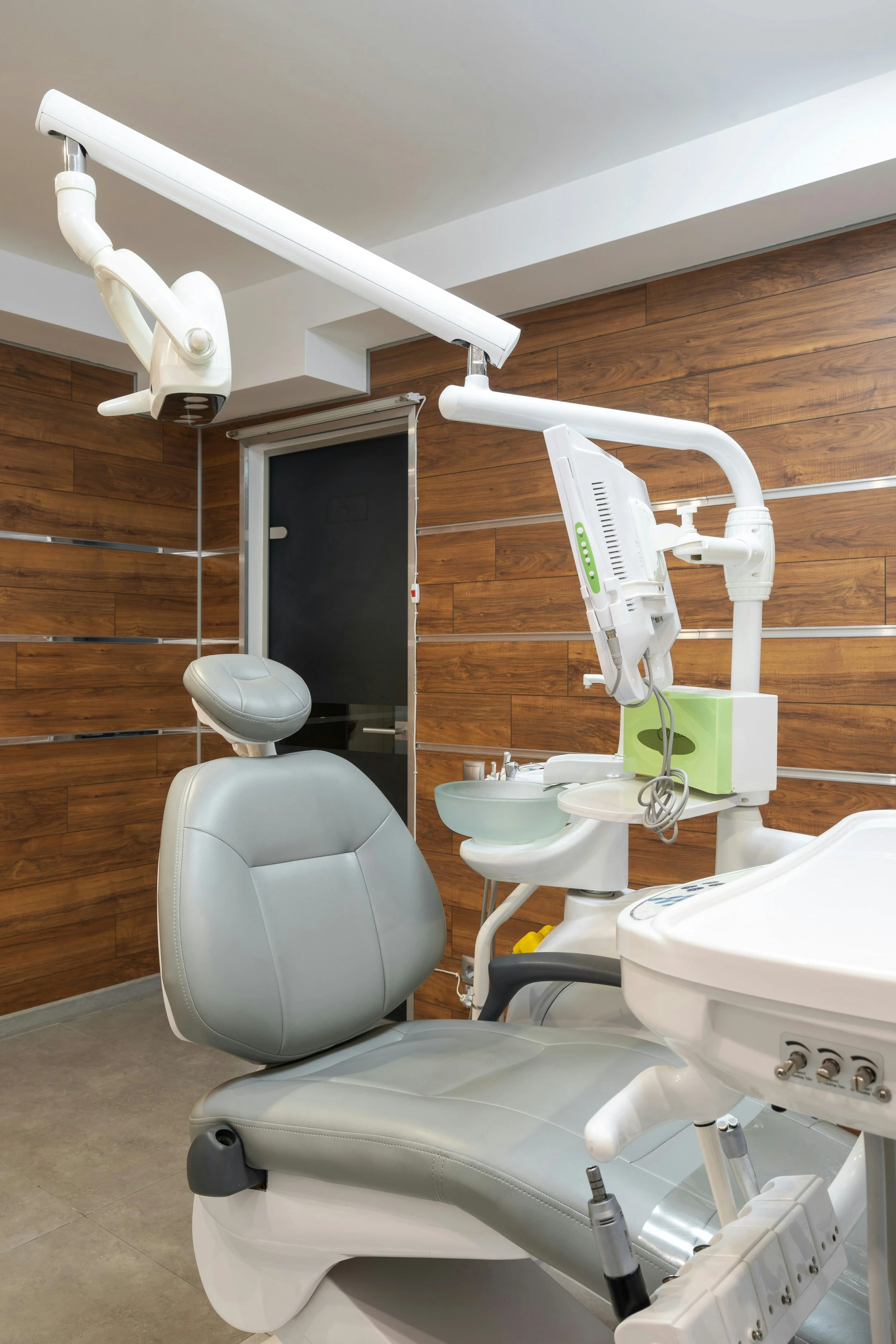 Dental examination room with a dentist chair, overhead light, and dental equipment against wood paneled wall.