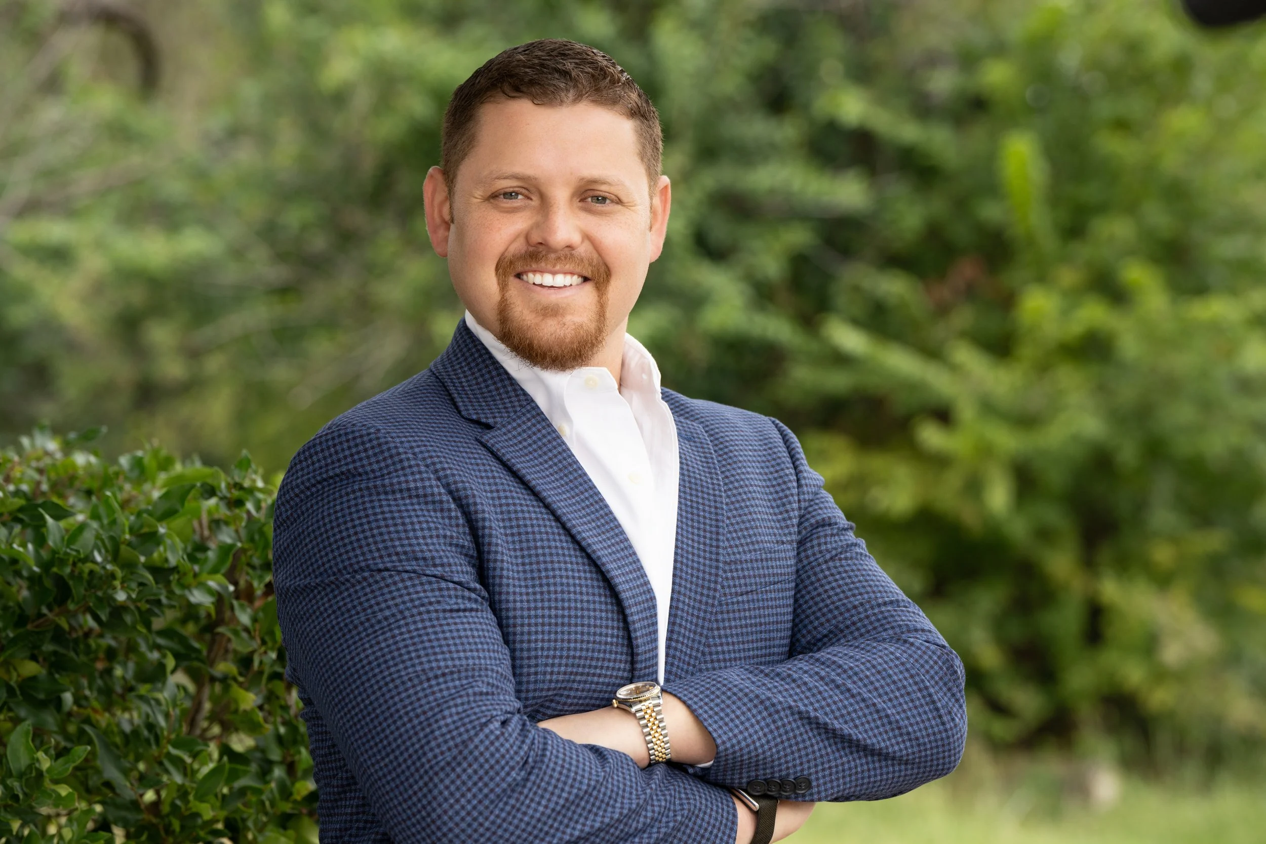 A man with short brown hair and a beard smiling, wearing a white shirt, a blue checkered blazer, and a wristwatch, standing outdoors with green foliage in the background.
