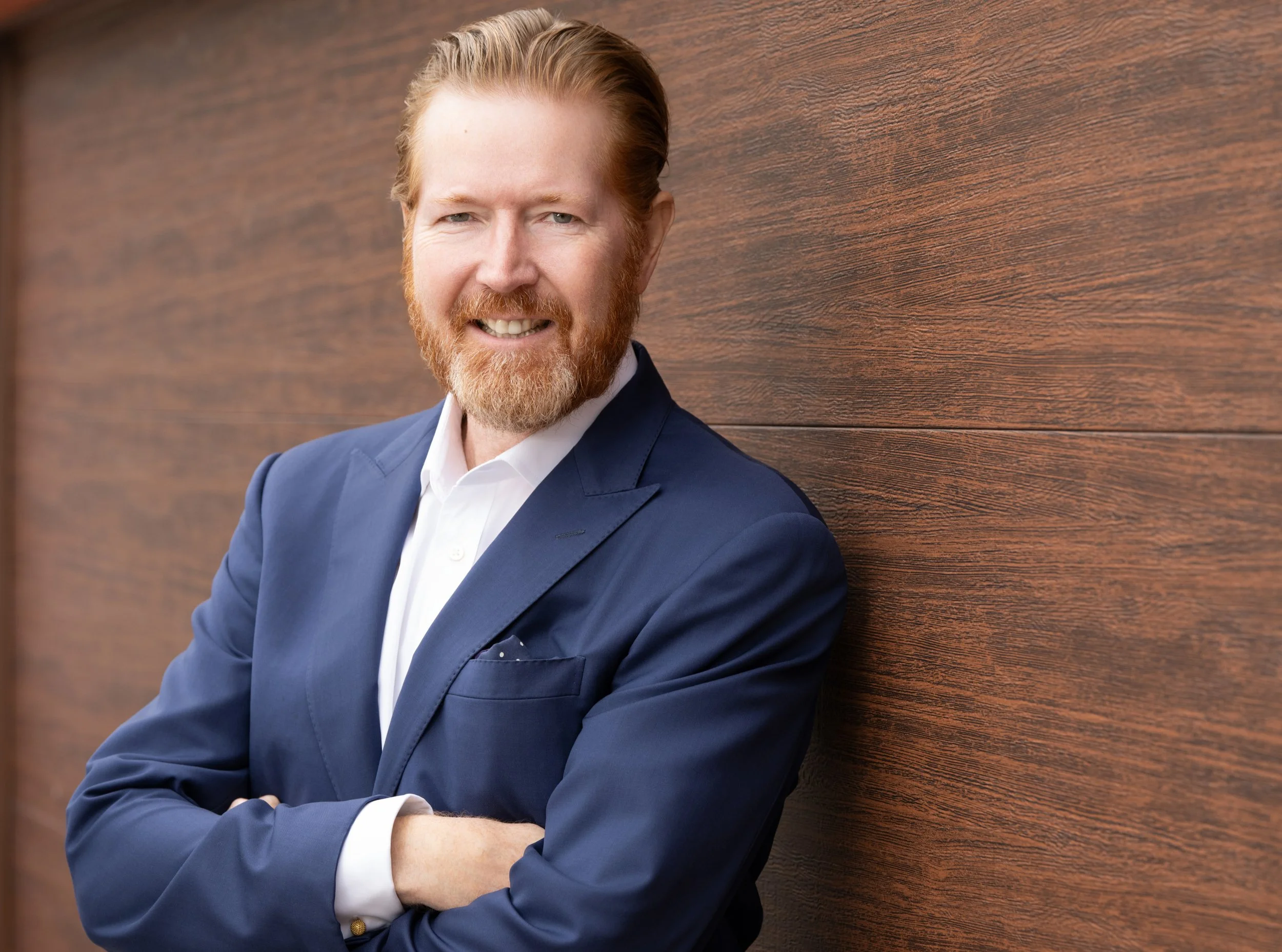 A man with red hair, beard, and wearing a dark blue suit and white shirt, standing against a wooden wall, smiling with arms crossed.