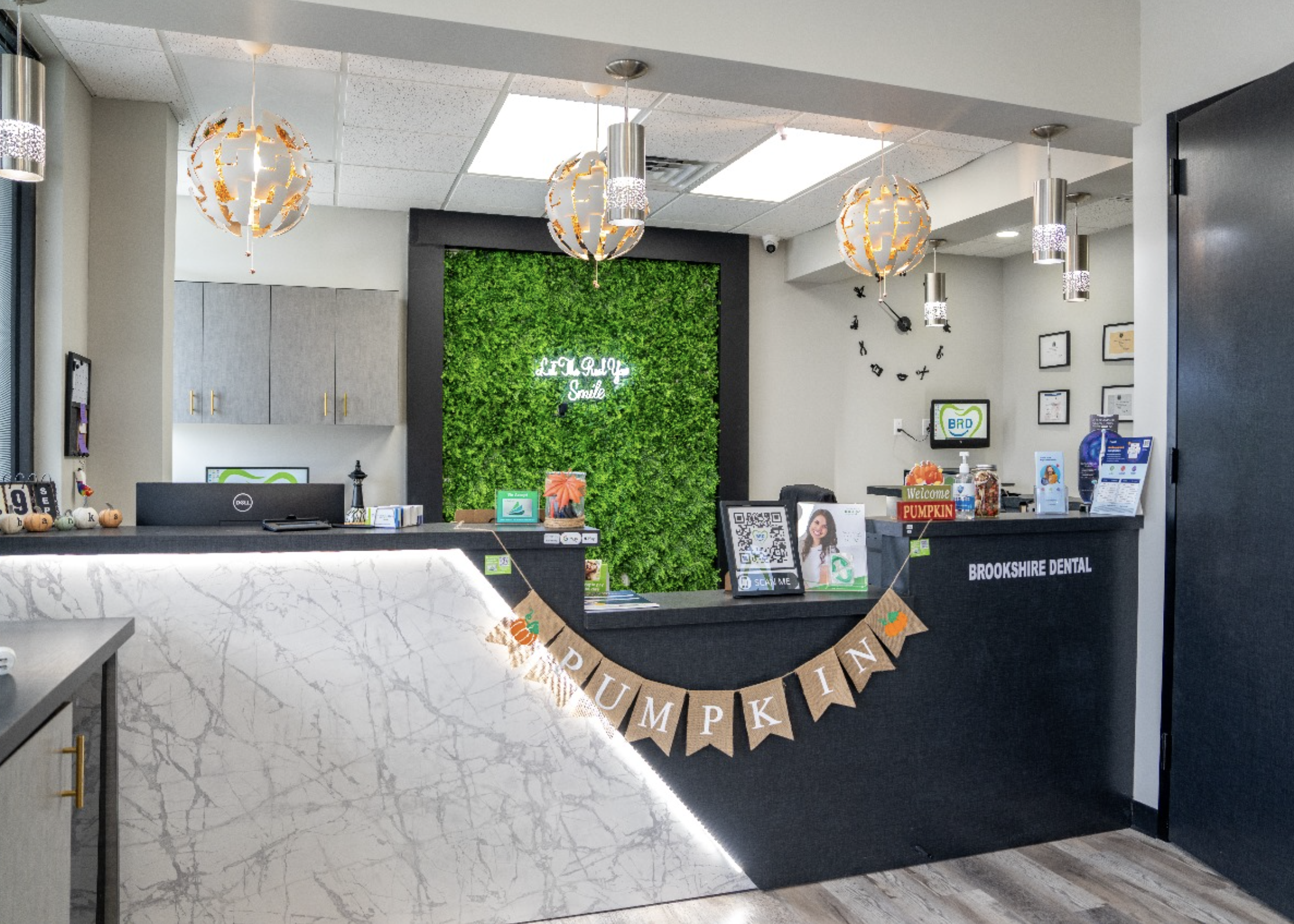 Reception area of a dental office decorated for autumn with pumpkin decorations. The front desk has a black counter with a 'Pumpkin' banner hanging. Behind the desk, there is a wall covered with green foliage and a neon sign that reads 'Smile.' The ceiling has decorative hanging lights and a large clock on the wall.