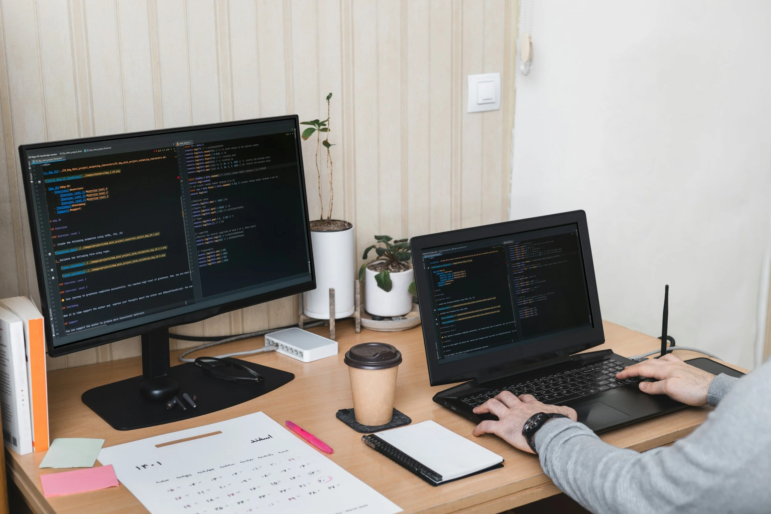 Person working on a laptop with coding on a dual monitor setup, on a wooden desk with plants, a coffee cup, a calendar, sticky notes, and writing materials.