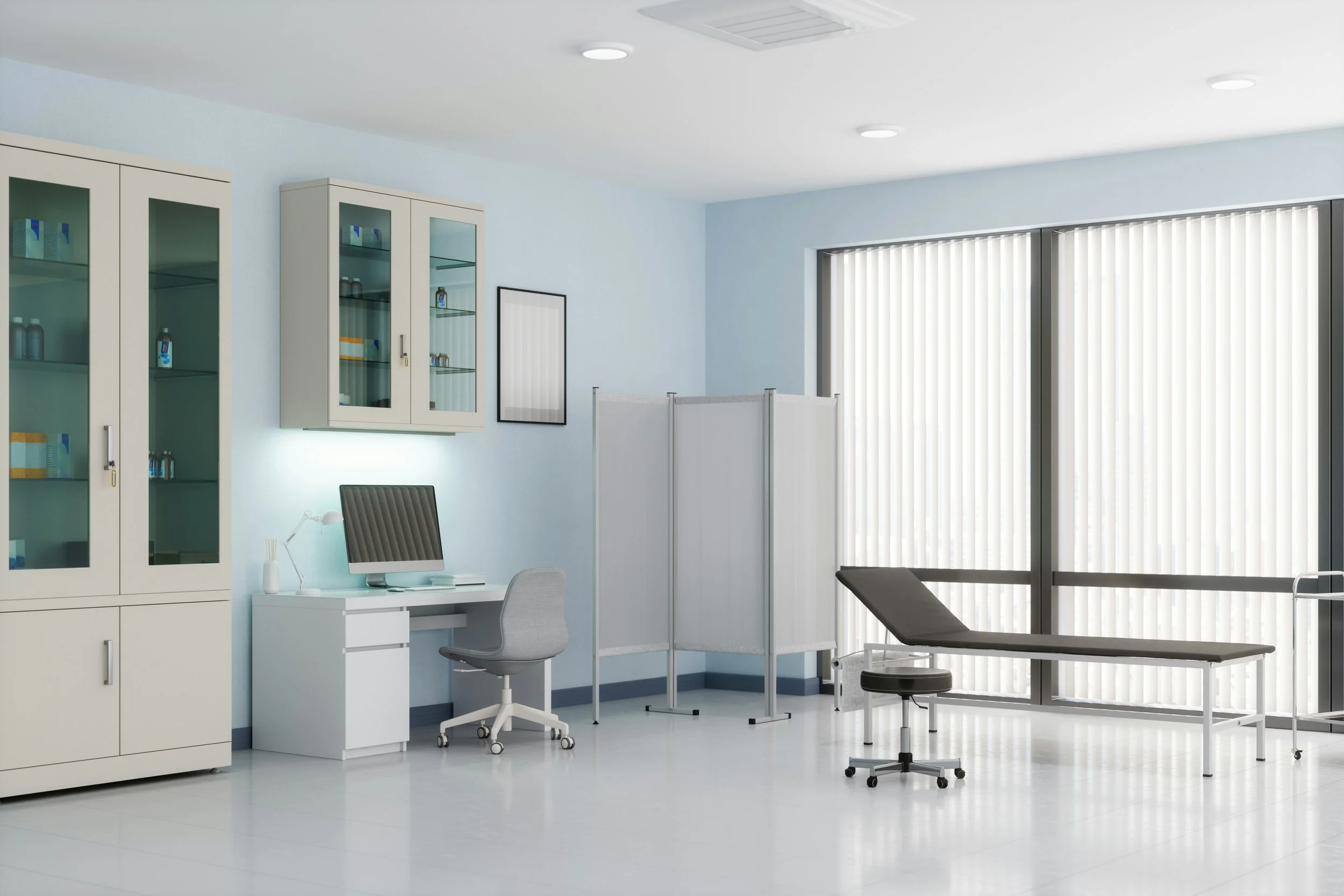 A medical examination room with a white examination bed near large windows with vertical blinds, medical storage cabinets, a computer on a white desk, a gray swivel chair, and a privacy divider.