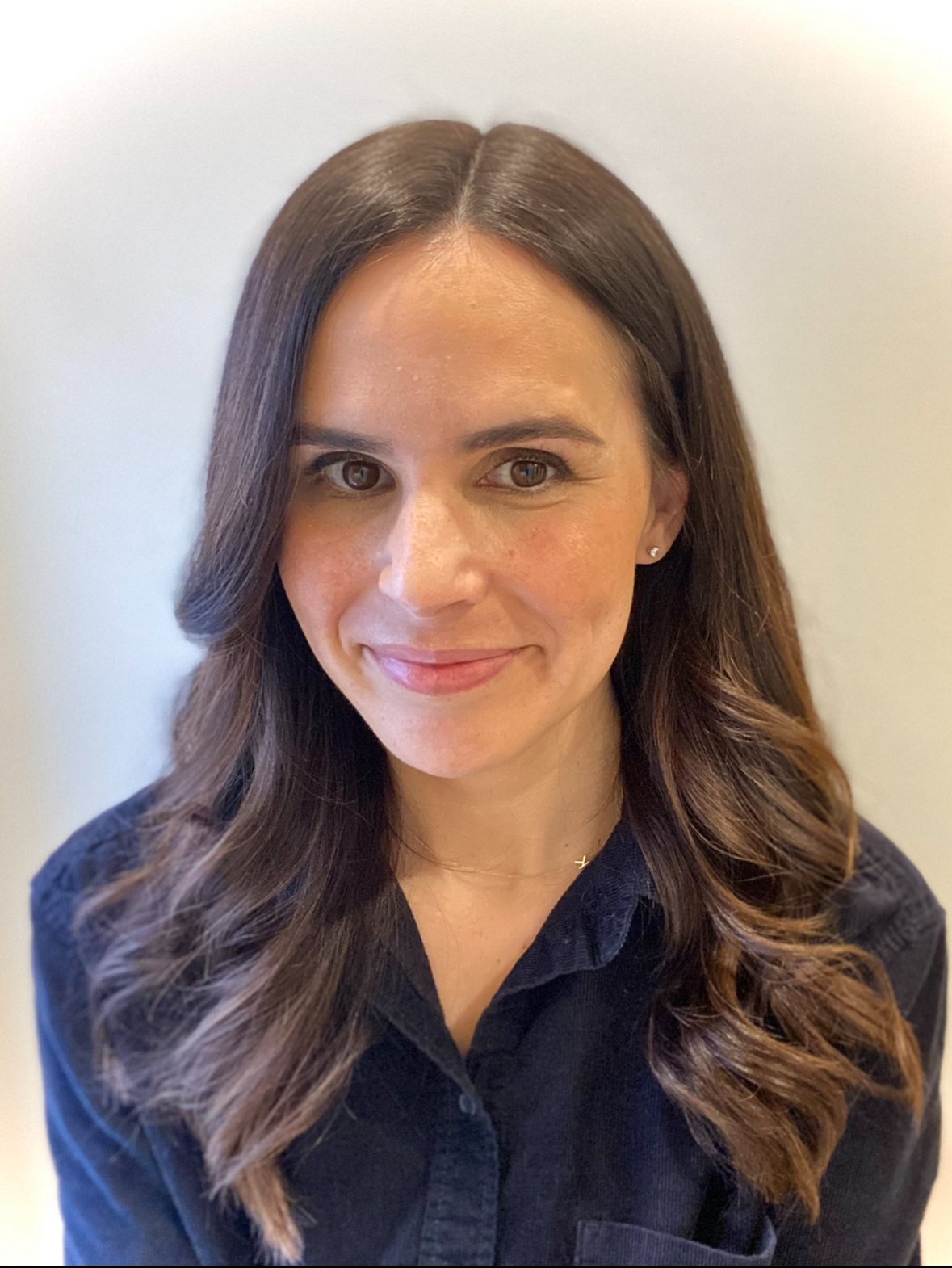 A portrait of a woman with long wavy brown hair, wearing a dark shirt, smiling softly, with a neutral background.