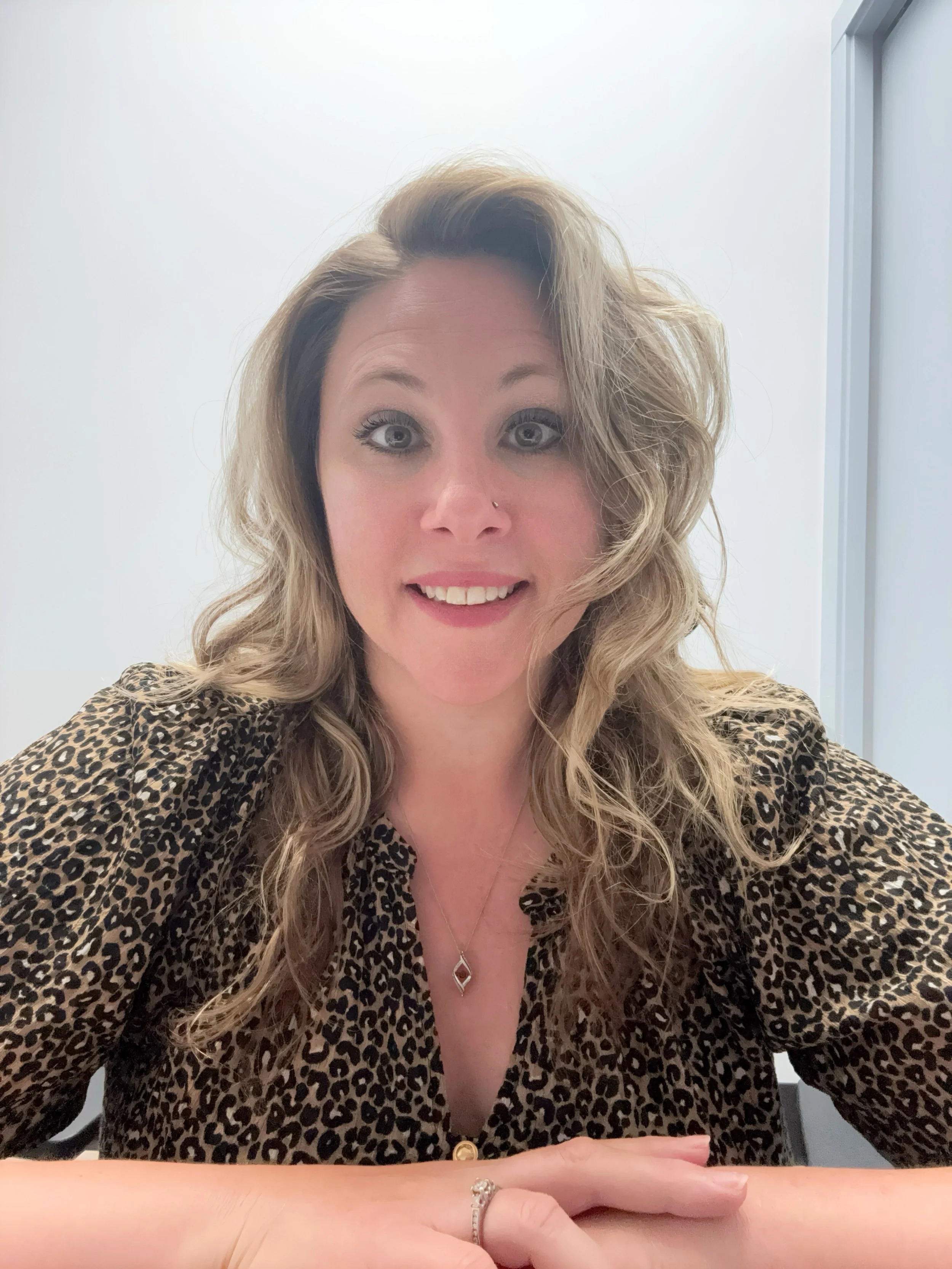 Close-up of a woman with blonde, wavy hair, wearing a leopard print shirt and jewelry, smiling at the camera in a well-lit room.