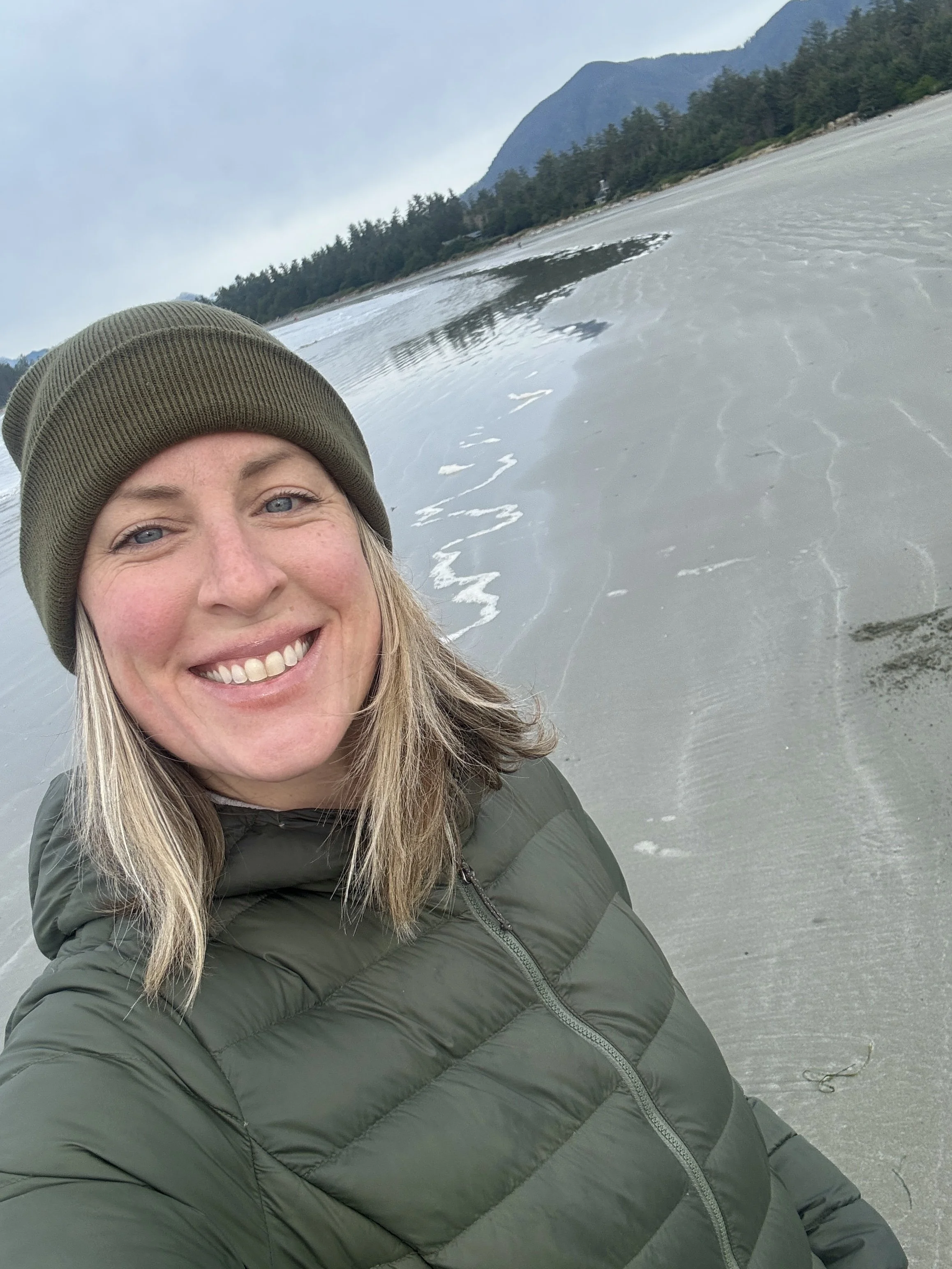 A woman smiling in a green jacket and knit cap standing on a beach with water and mountains in the background.