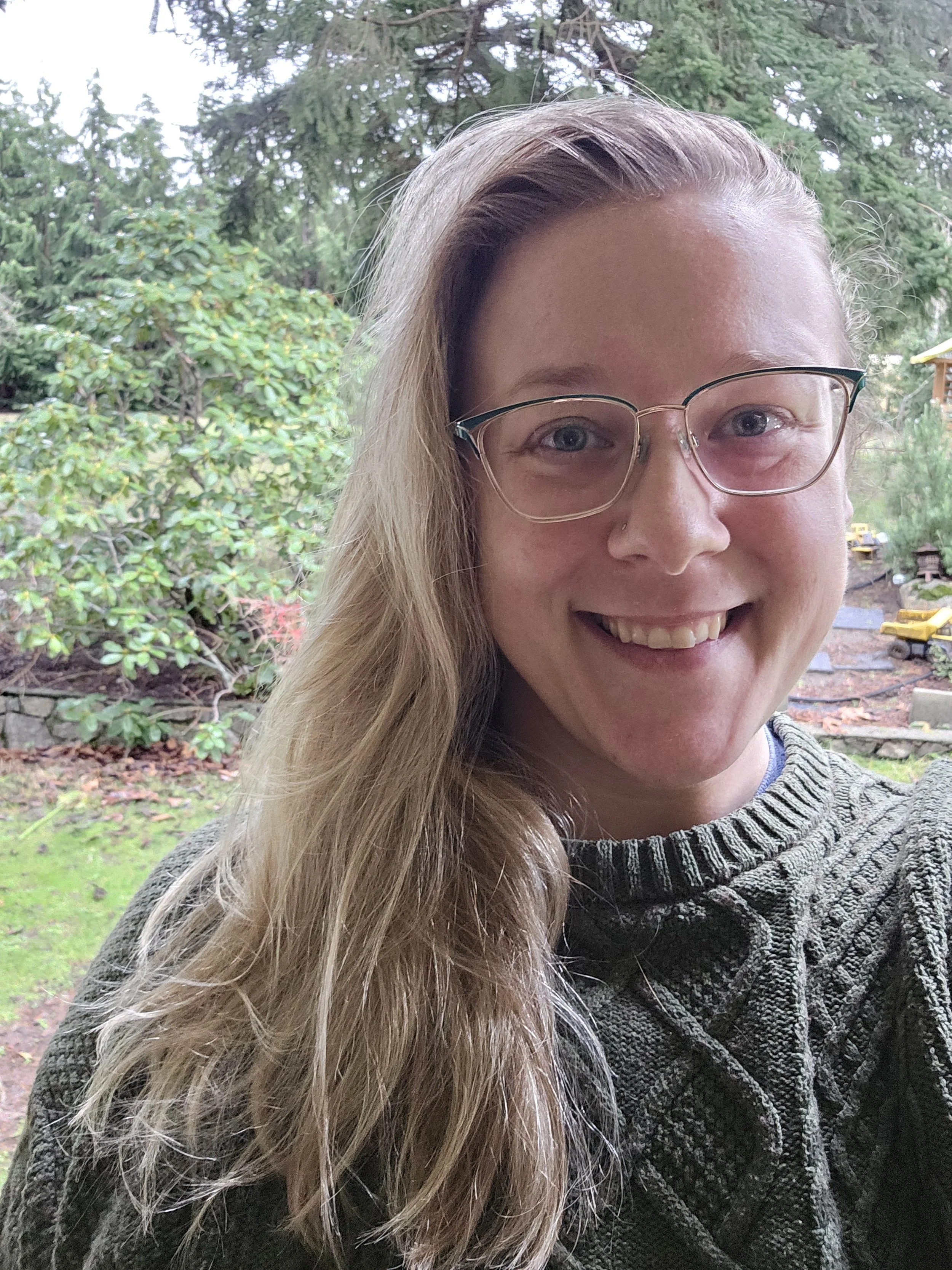 A smiling woman with glasses and long blonde hair taking a selfie outdoors in a lush, green garden.