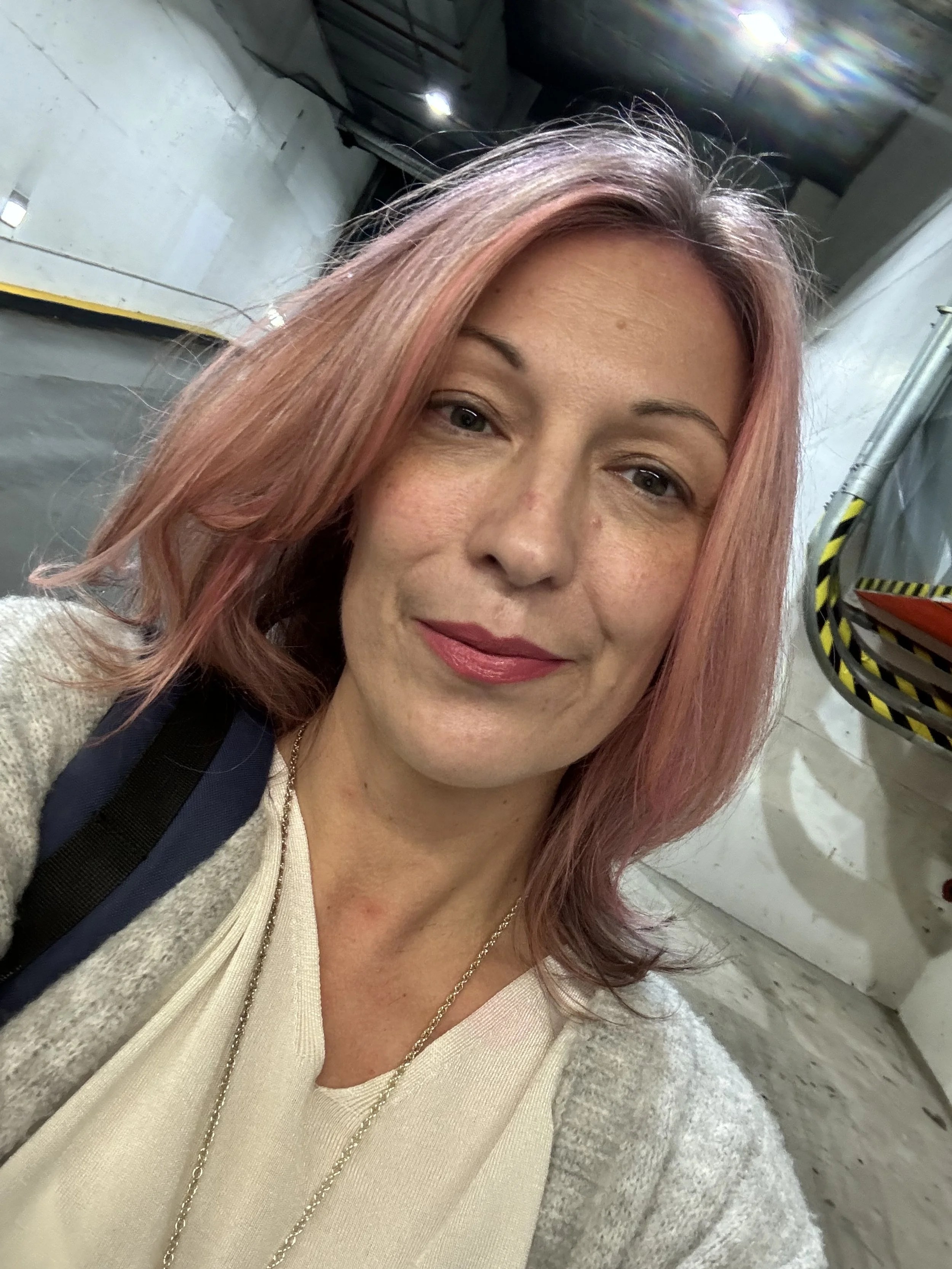 A woman with pinkish hair and light skin taking a selfie in an industrial or warehouse setting. She is smiling softly, wearing a beige top, a gray sweater, and a blue backpack strap is visible on her shoulder.