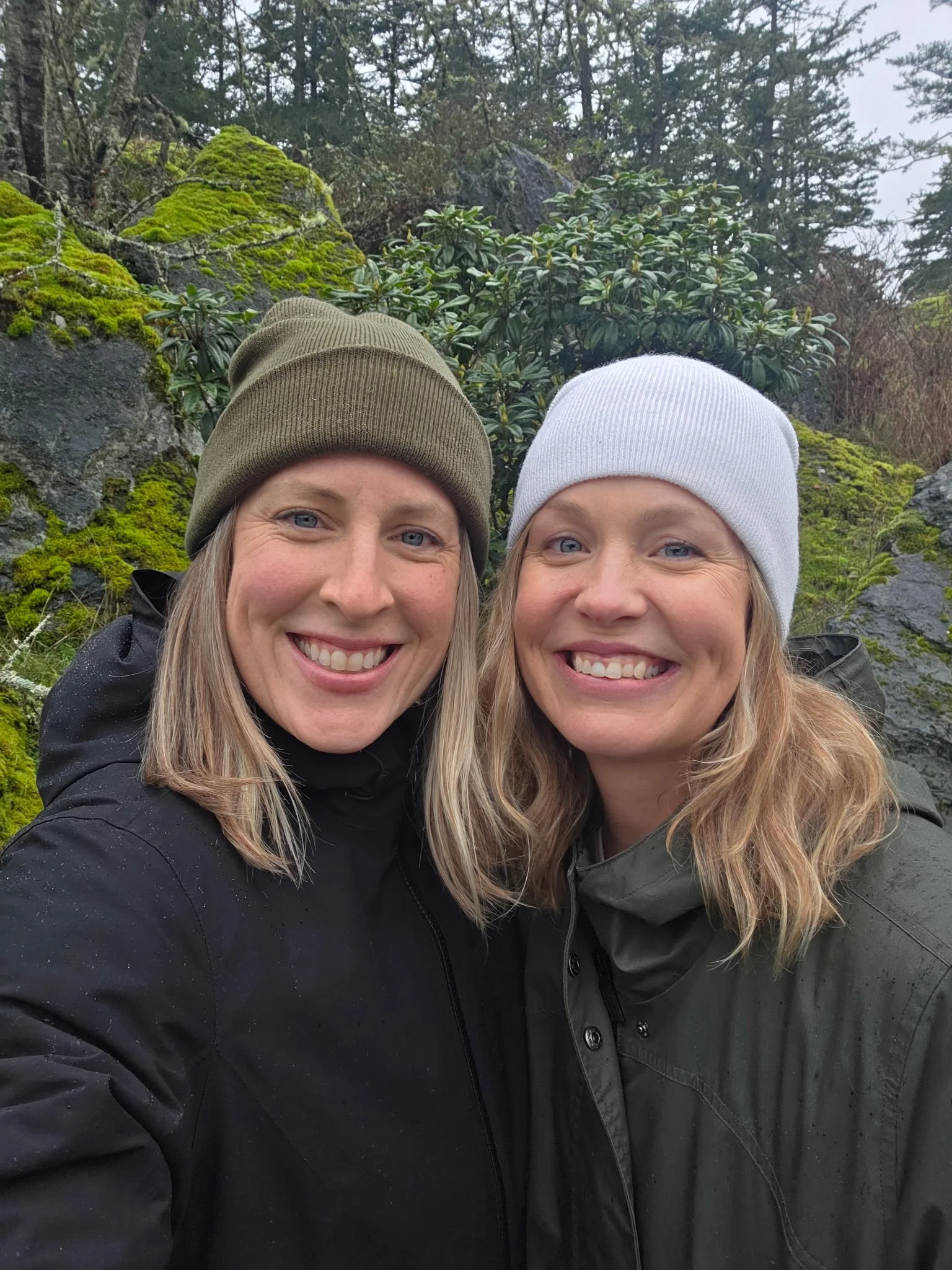 Two women smiling outdoors, wearing jackets and knit hats, standing on a moss-covered rocky terrain with trees and vegetation in the background.