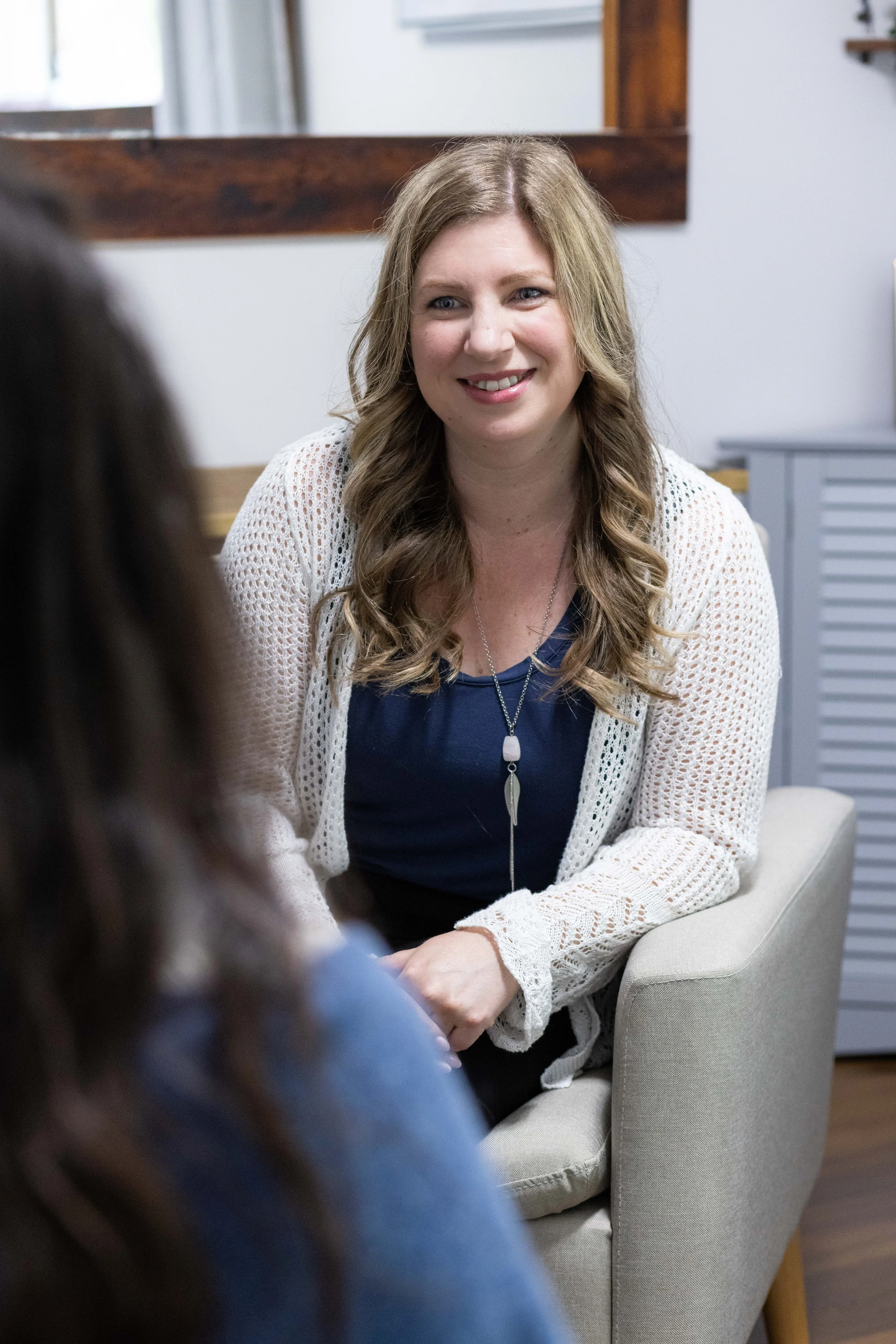 A woman with long wavy blond hair smiling while seated and talking to a person whose back is visible, in an indoor setting.