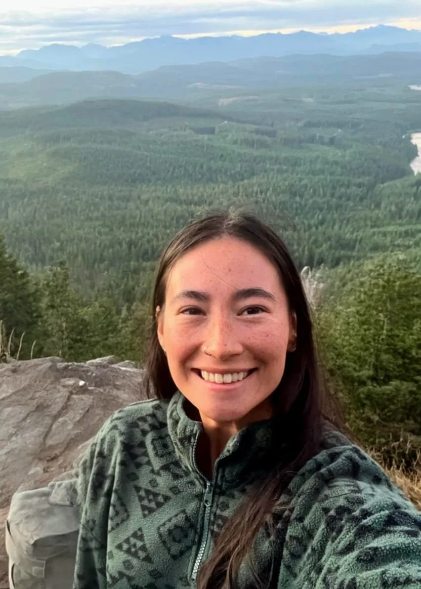 A woman taking a selfie on a mountain with a scenic view of forests, rolling hills, and distant mountains under a cloudy sky.