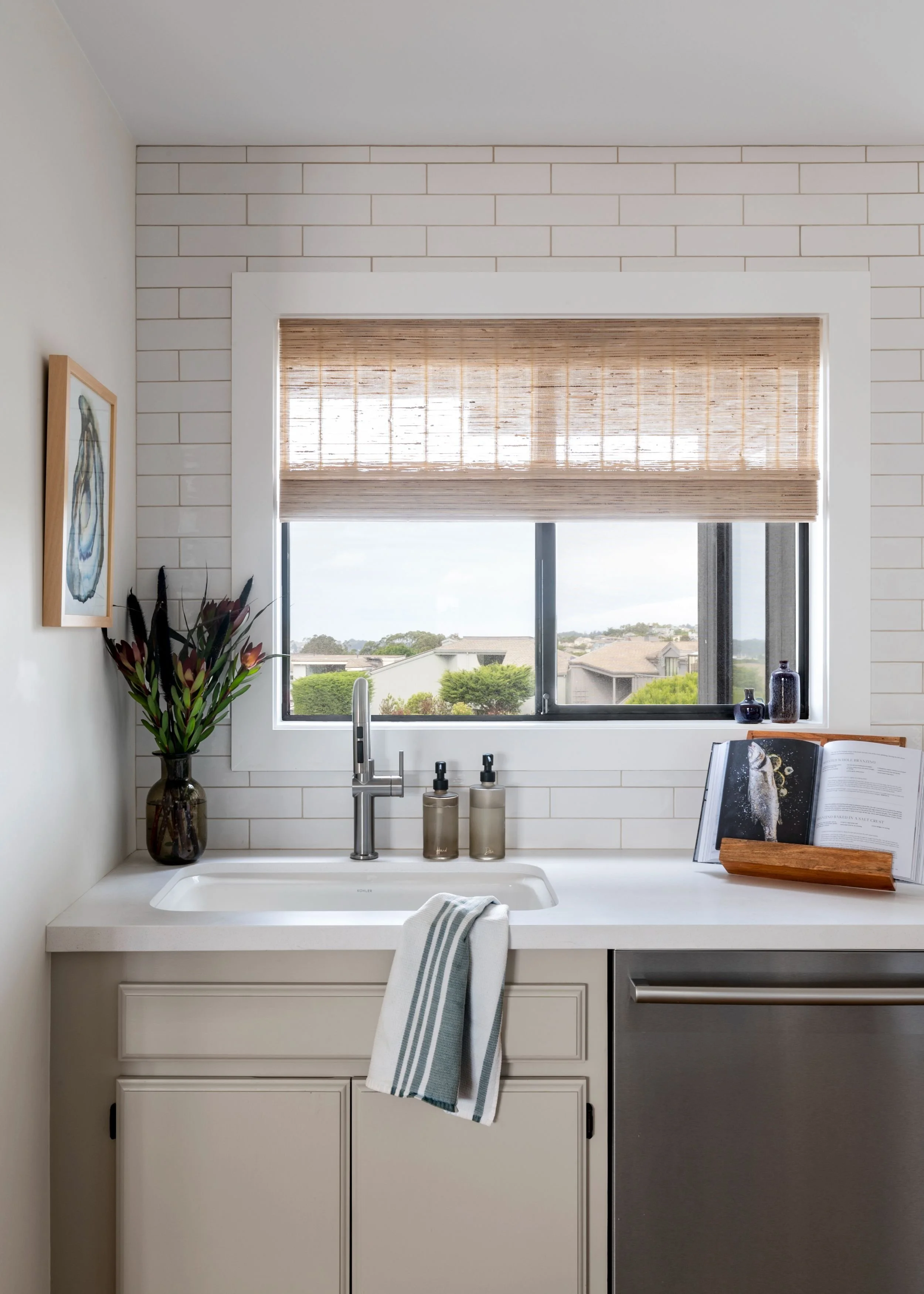 A kitchen sink with a window above it, decorated with a potted plant, soap dispensers, and an open cookbook on a wooden stand.