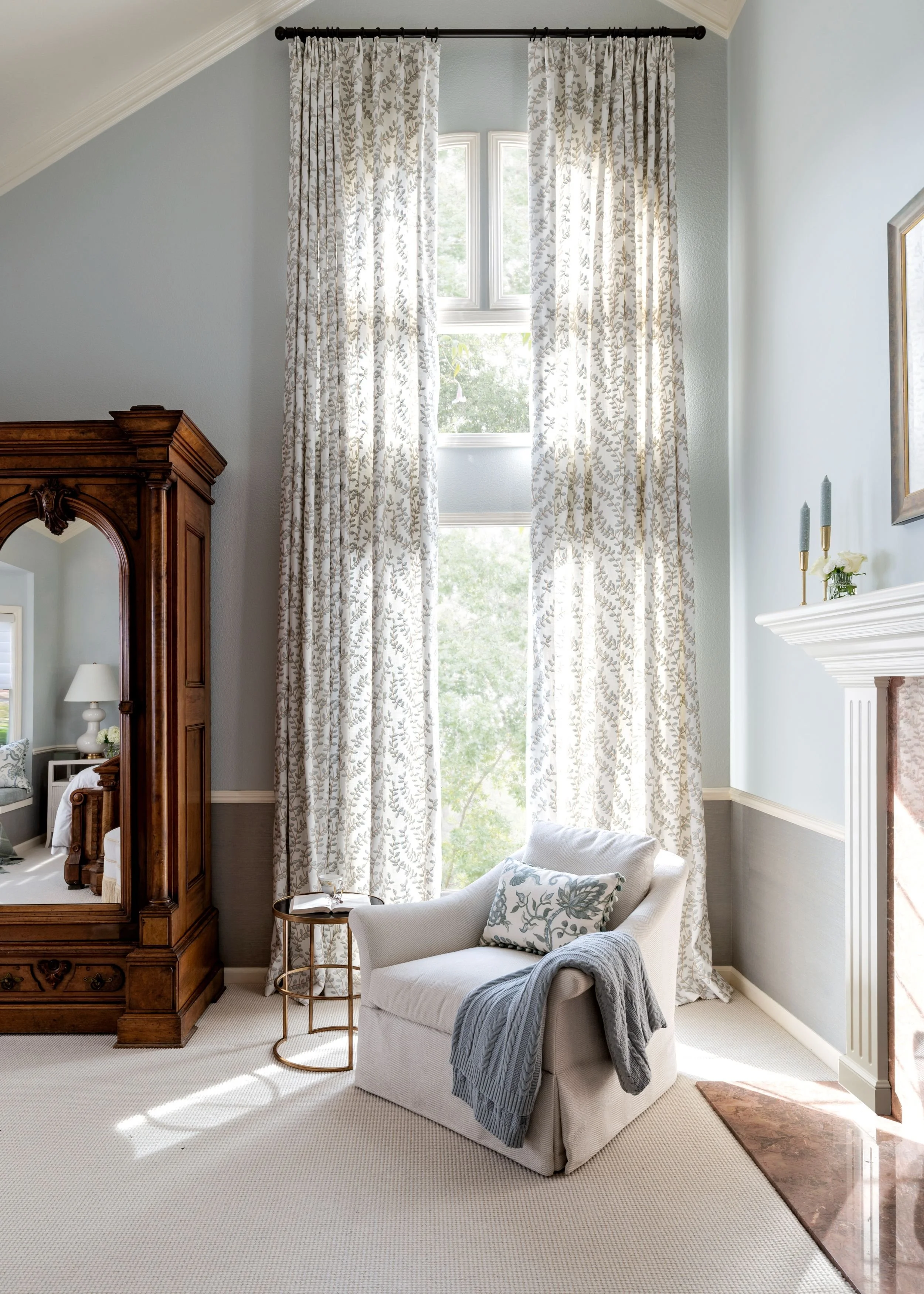 A cozy corner of a room with a white armchair, a pillow, and a blanket, next to a window with long floral curtains. There is a small side table with a book, an armoire with a mirror, a fireplace with candles and a small plant, and a view of trees out