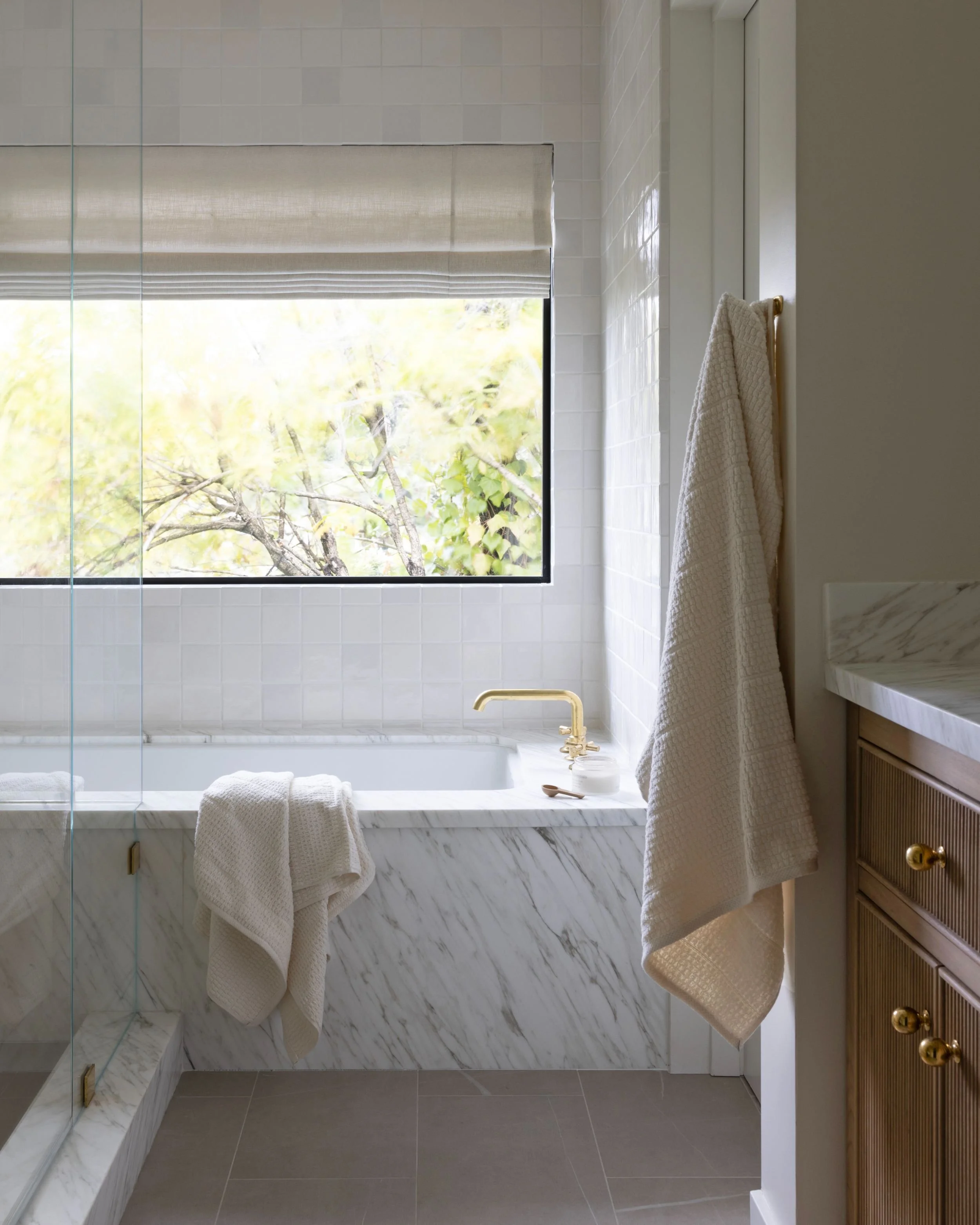 A bathroom with a large window, a marble bathtub, brass faucet, and beige towels hanging on a wooden cabinet.
