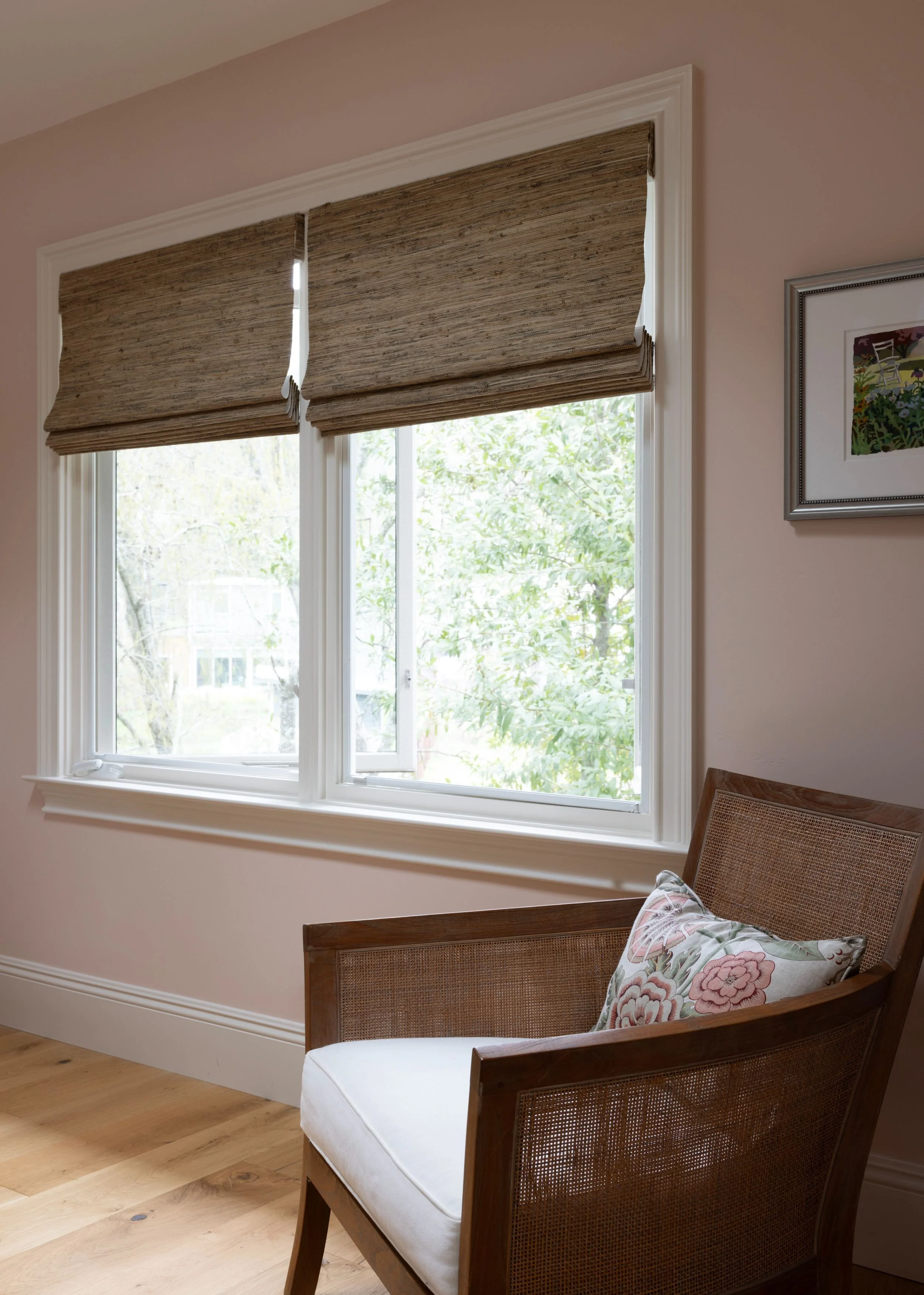 A cozy corner with a wooden armchair featuring a cushion and floral pillow, beside a large window with beige roller shades, overlooking greenery outside, and a framed picture on light pink wall.