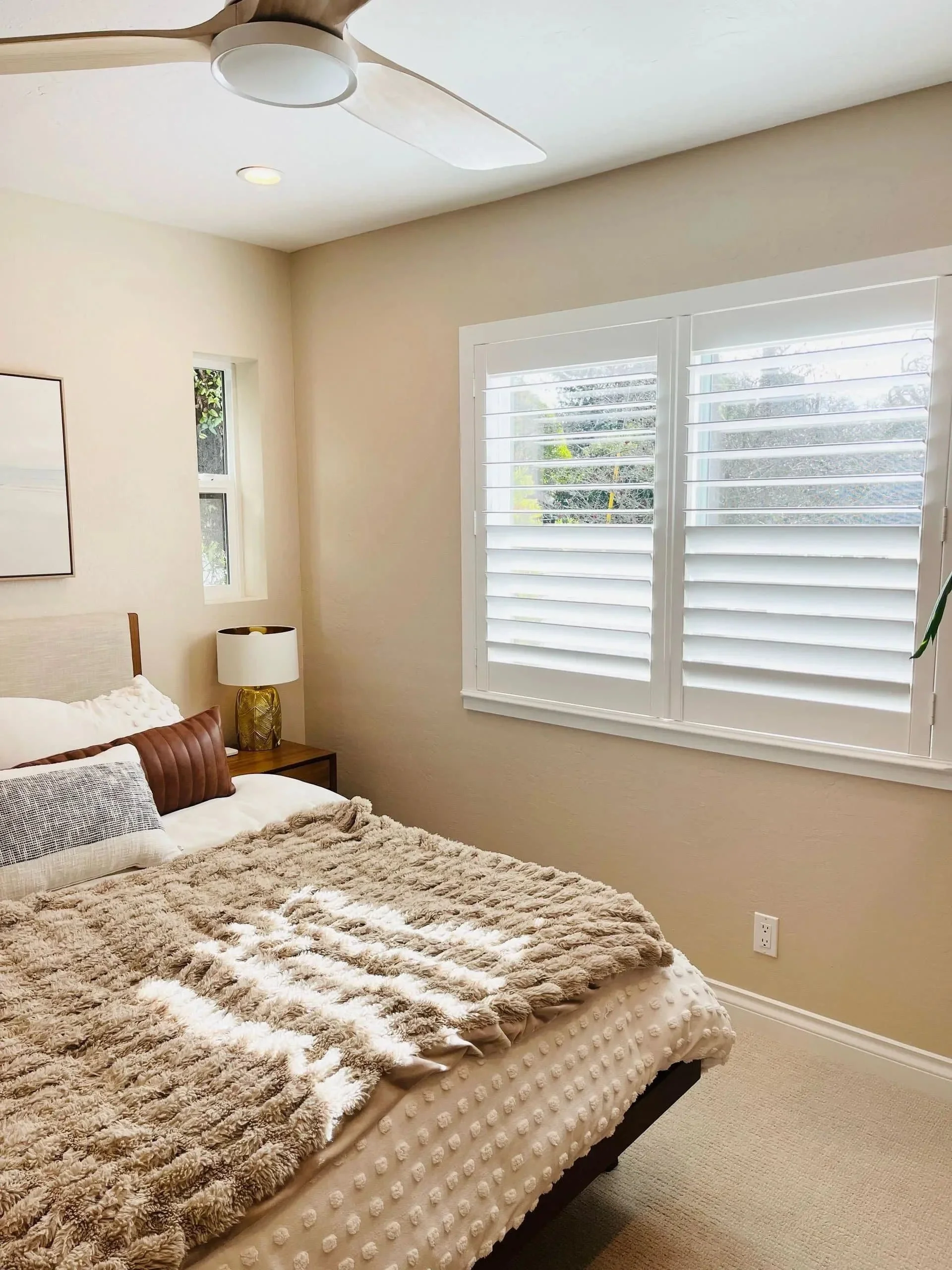A cozy bedroom featuring a bed with textured beige bedding and decorative pillows, a side table with a lamp, a ceiling fan, and a window with white plantation shutters allowing natural light.