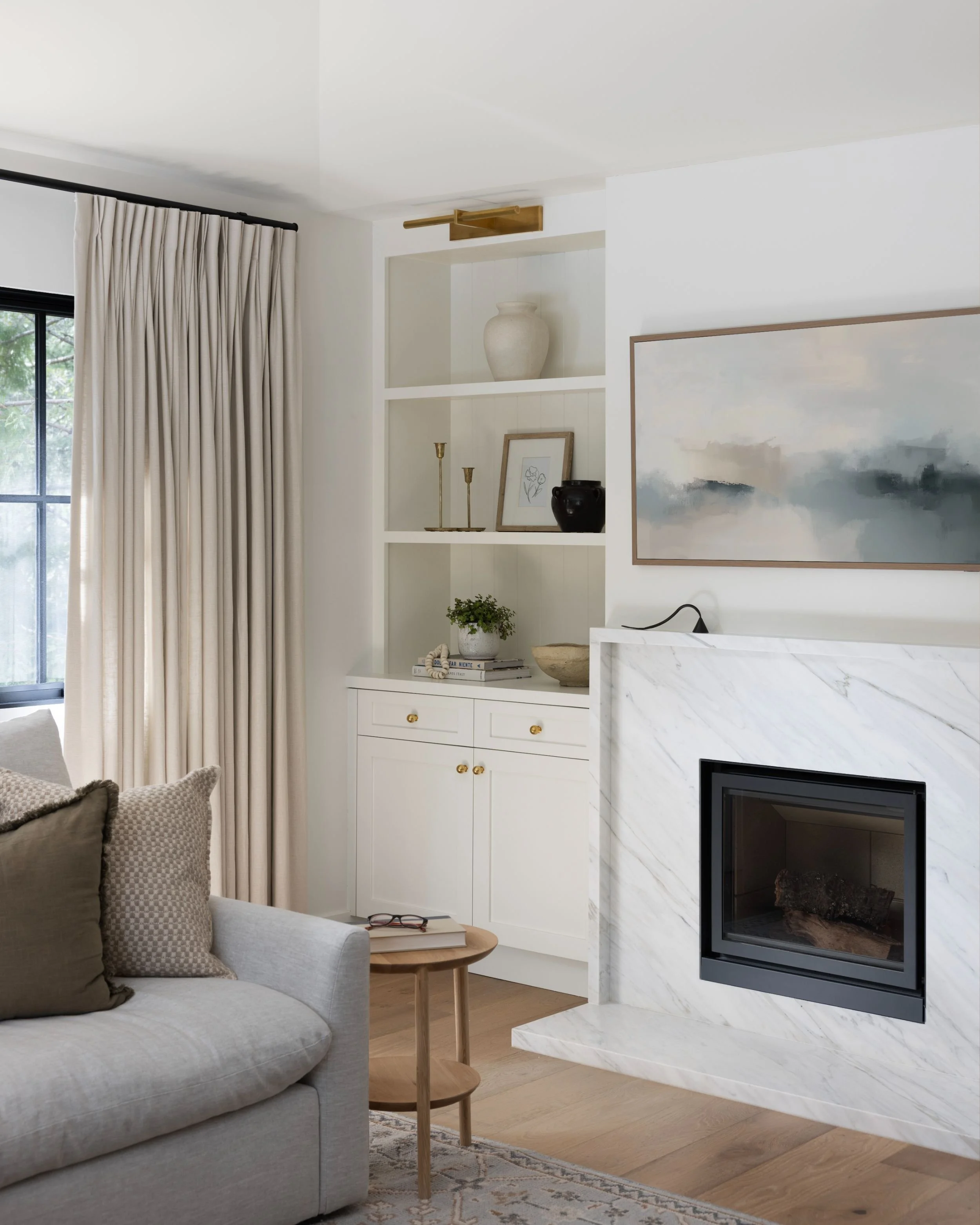 Living room with white walls, beige curtains, a gray sofa with beige and brown pillows, a wooden side table, a white built-in cabinet with decorative objects on top, and a marble fireplace with a painting above it.
