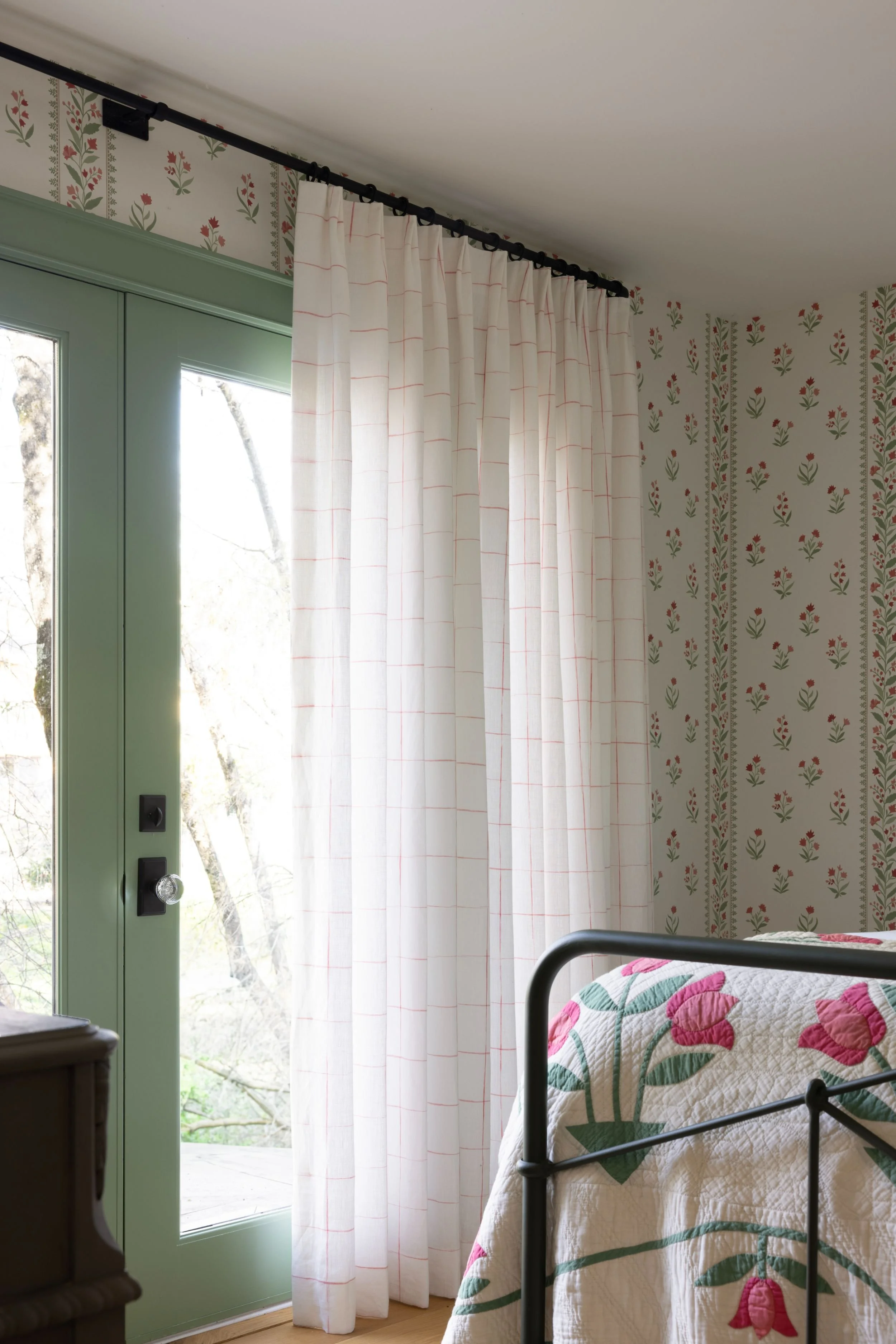 A bedroom with floral wallpaper, a bed with a quilted blanket featuring pink flowers and green leaves, white curtains with a faint pink grid, and a green sliding glass door with a handle and lock.