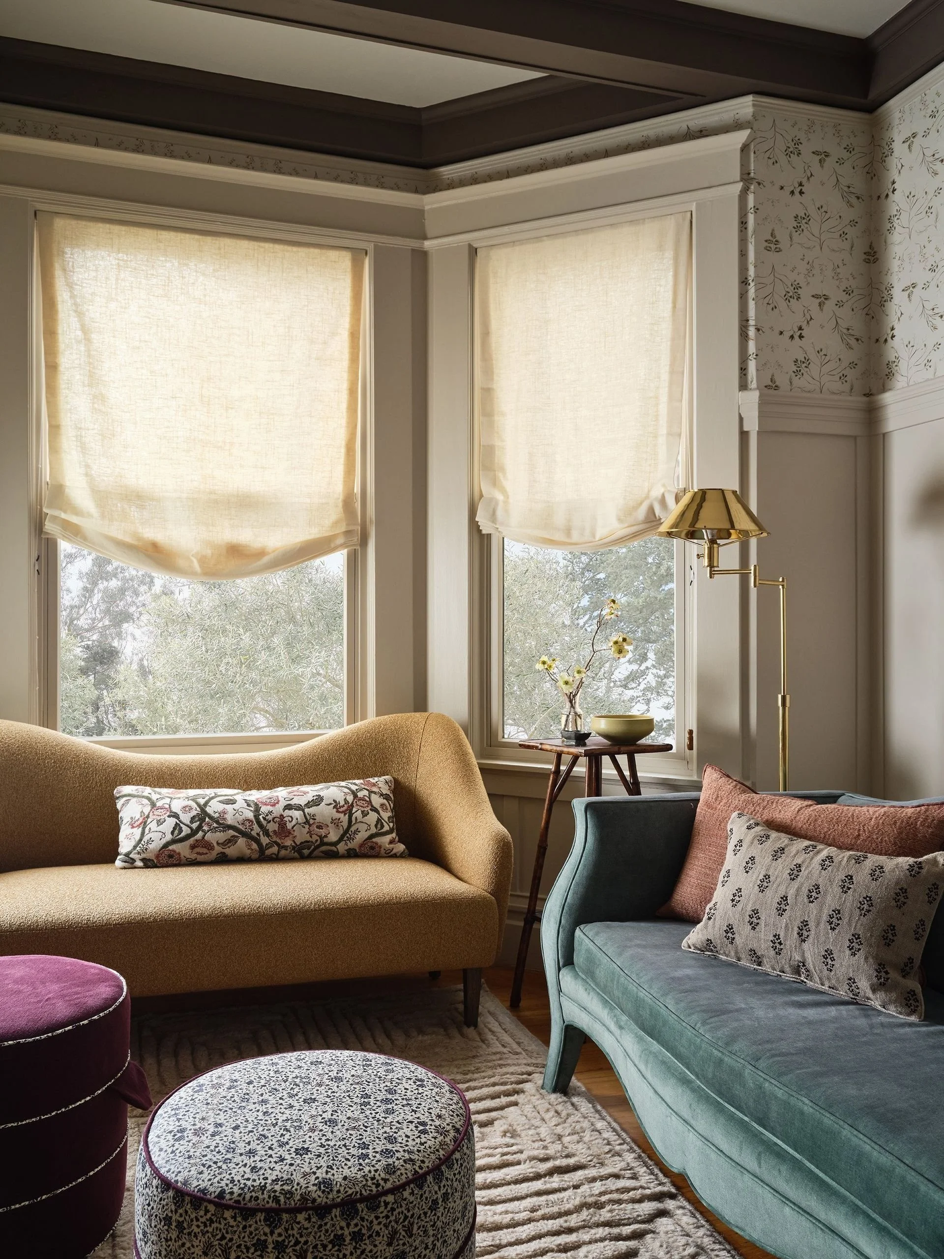 A cozy living room with two large windows covered by beige Roman shades, a mustard-colored sofa with a decorative pillow, a teal velvet armchair with pillows, a wooden side table with a flower vase, a gold floor lamp, a patterned area rug, and a purp