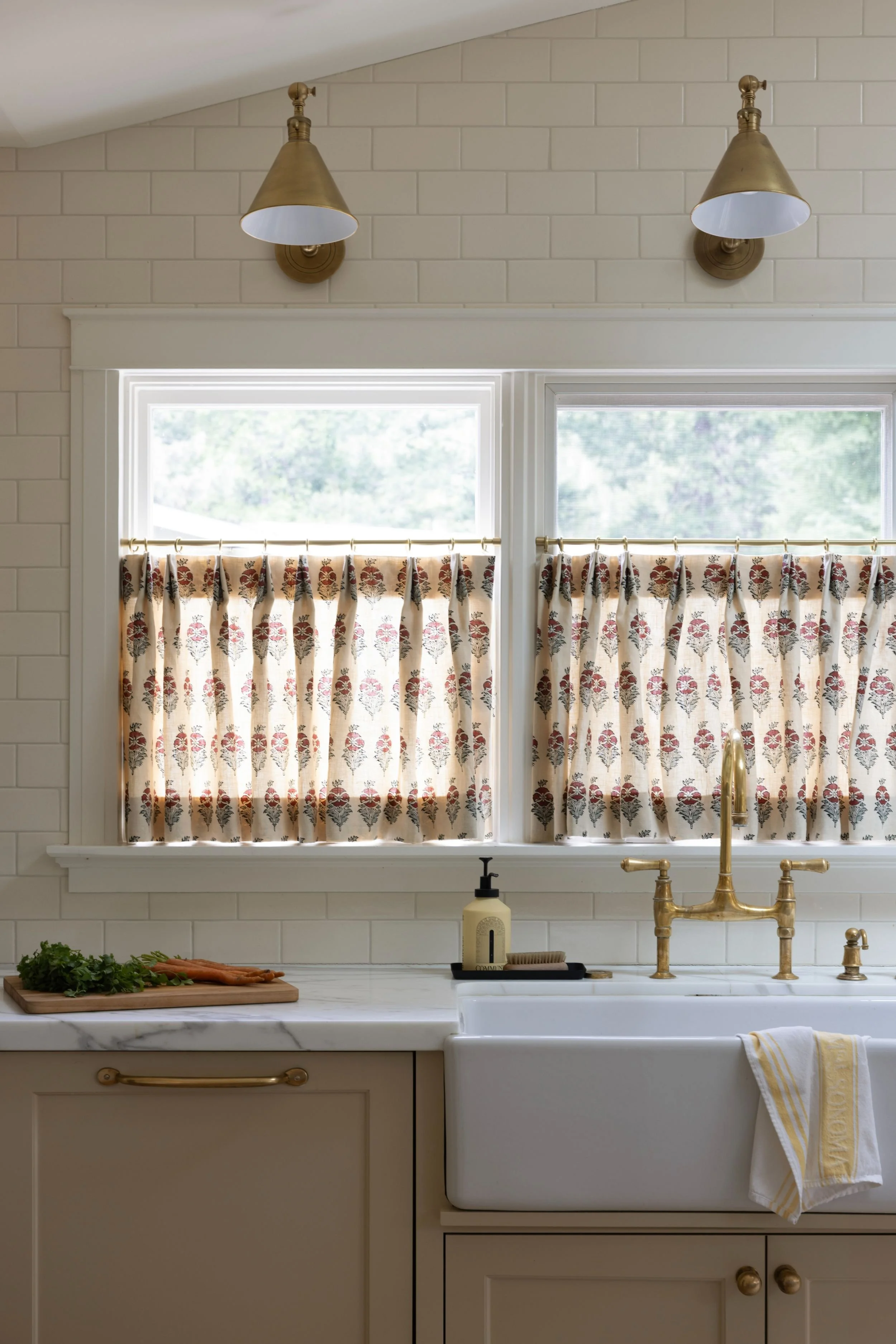 A kitchen with two large windows covered by floral curtains, a white farmhouse sink with a gold faucet, a beige cabinet, a marble countertop, and two wall-mounted gold lamps.
