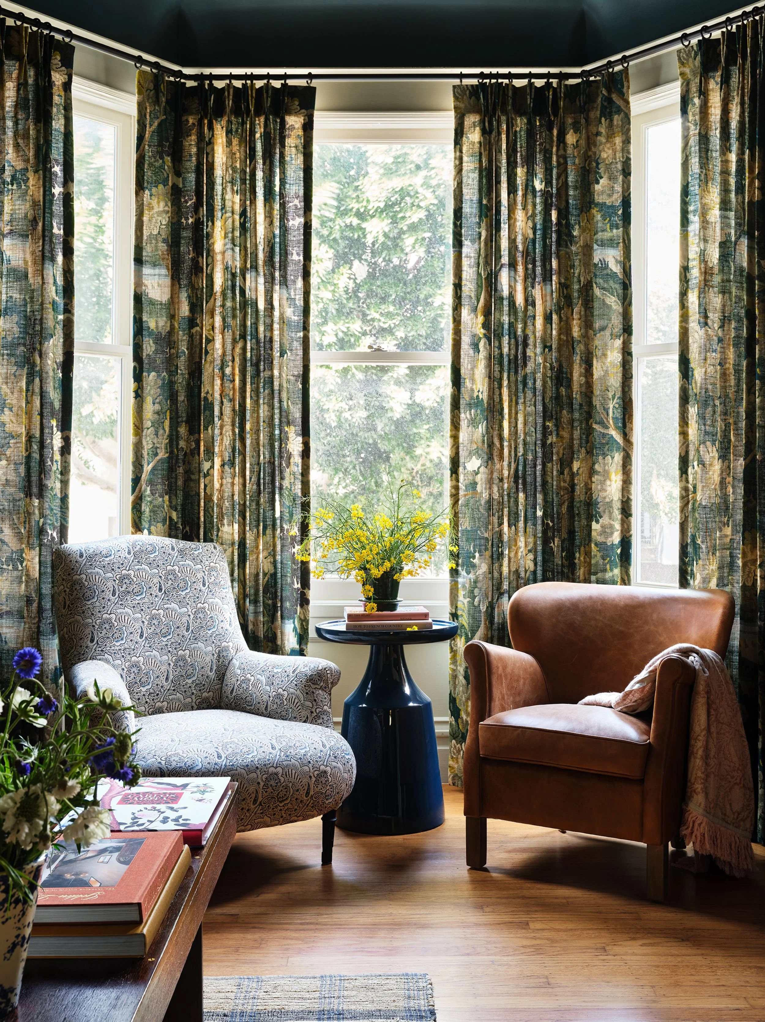 A cozy living room corner with two armchairs, one patterned and one leather, around a small black table with a vase of yellow flowers, in front of large windows with floral curtains, and a wooden floor with a small rug.