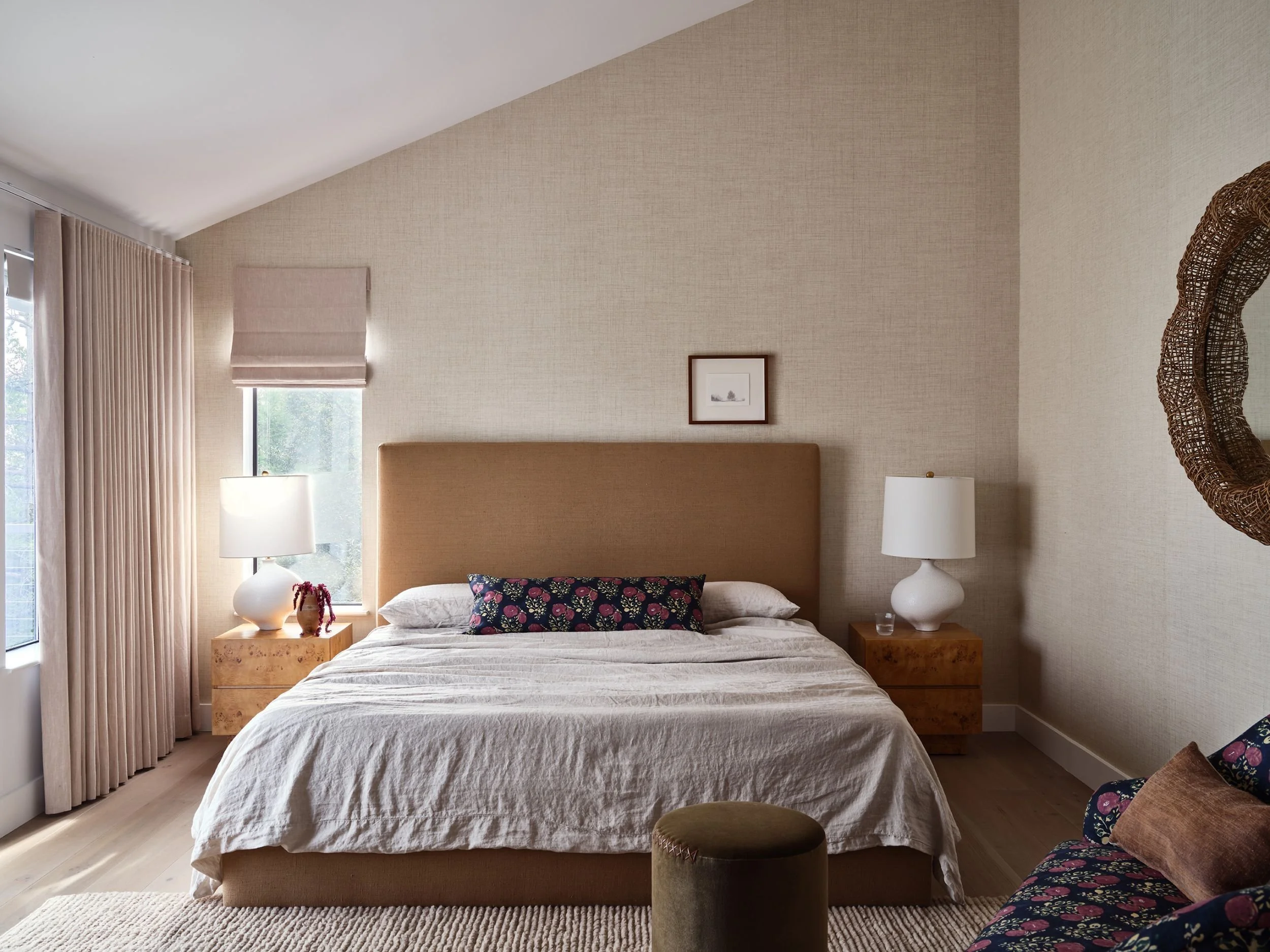 A cozy bedroom with a large bed, beige headboard, and white bedding. Flanking the bed are nightstands with white lamps, and a small framed picture hangs above the headboard. There are windows with curtains on the left, and a floral-patterned chair wi