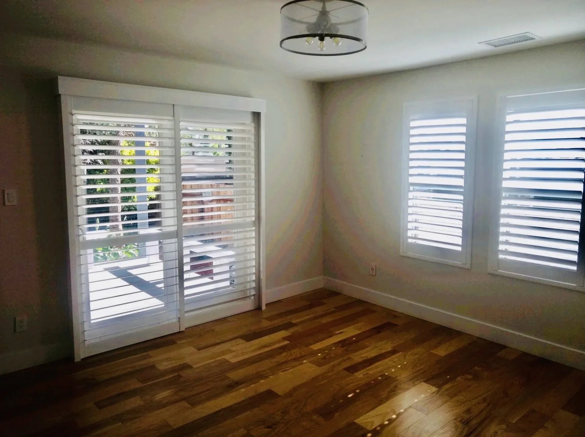Empty room with wooden floor, three large windows with white shutters, and a glass sliding door with matching shutters.