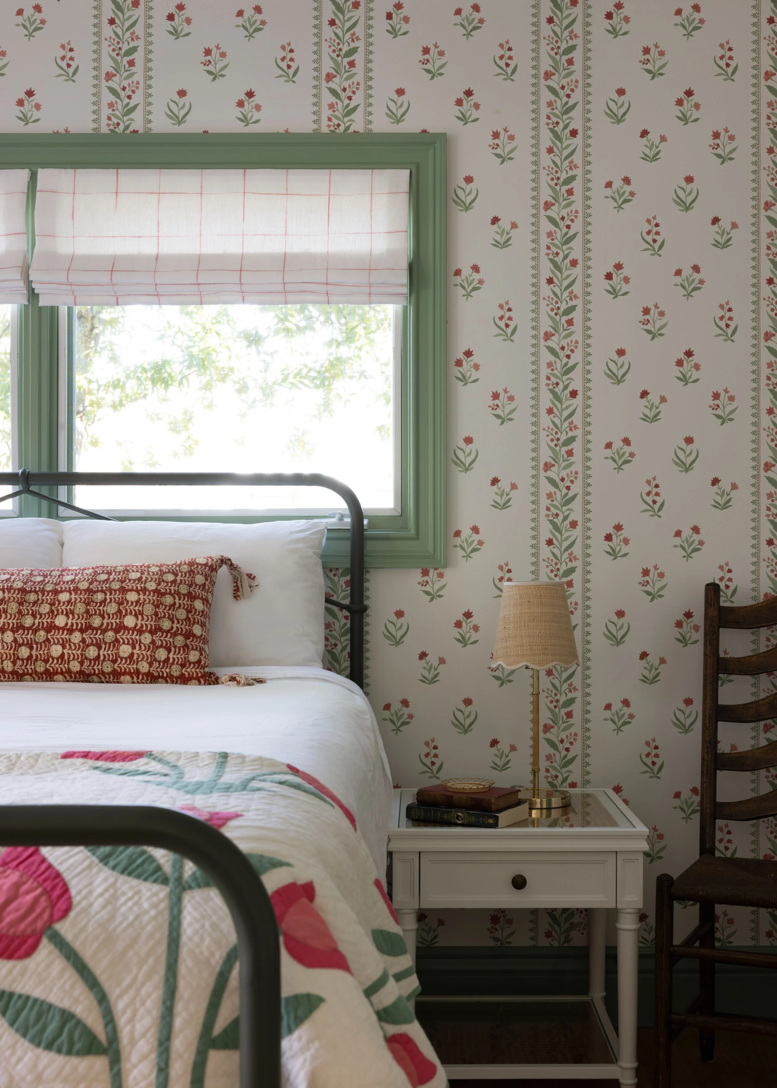 Cozy bedroom with floral wallpaper, a window with green trim and a white Roman shade, a bed with a patchwork quilt and red patterned pillow, a white bedside table with books and a beige lamp, and a wooden chair.