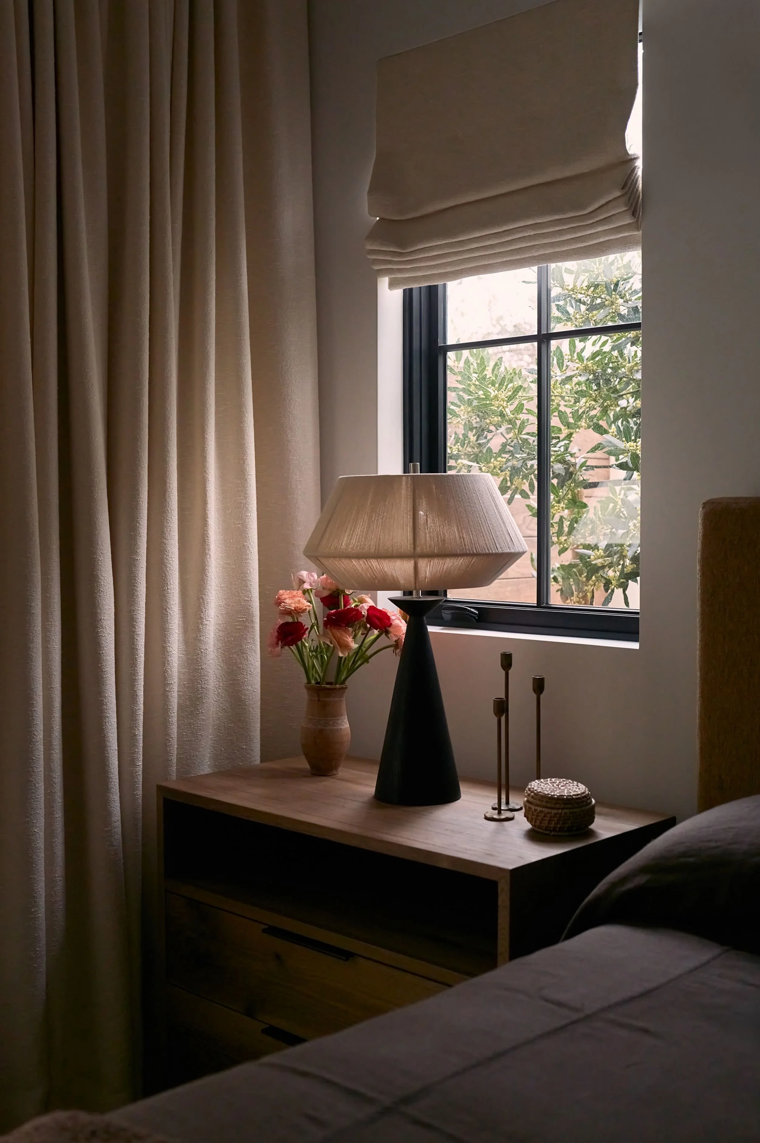 Nightstand with a flower vase, a table lamp, candles, and a decorative box near a window with curtains and a beige Roman shade.