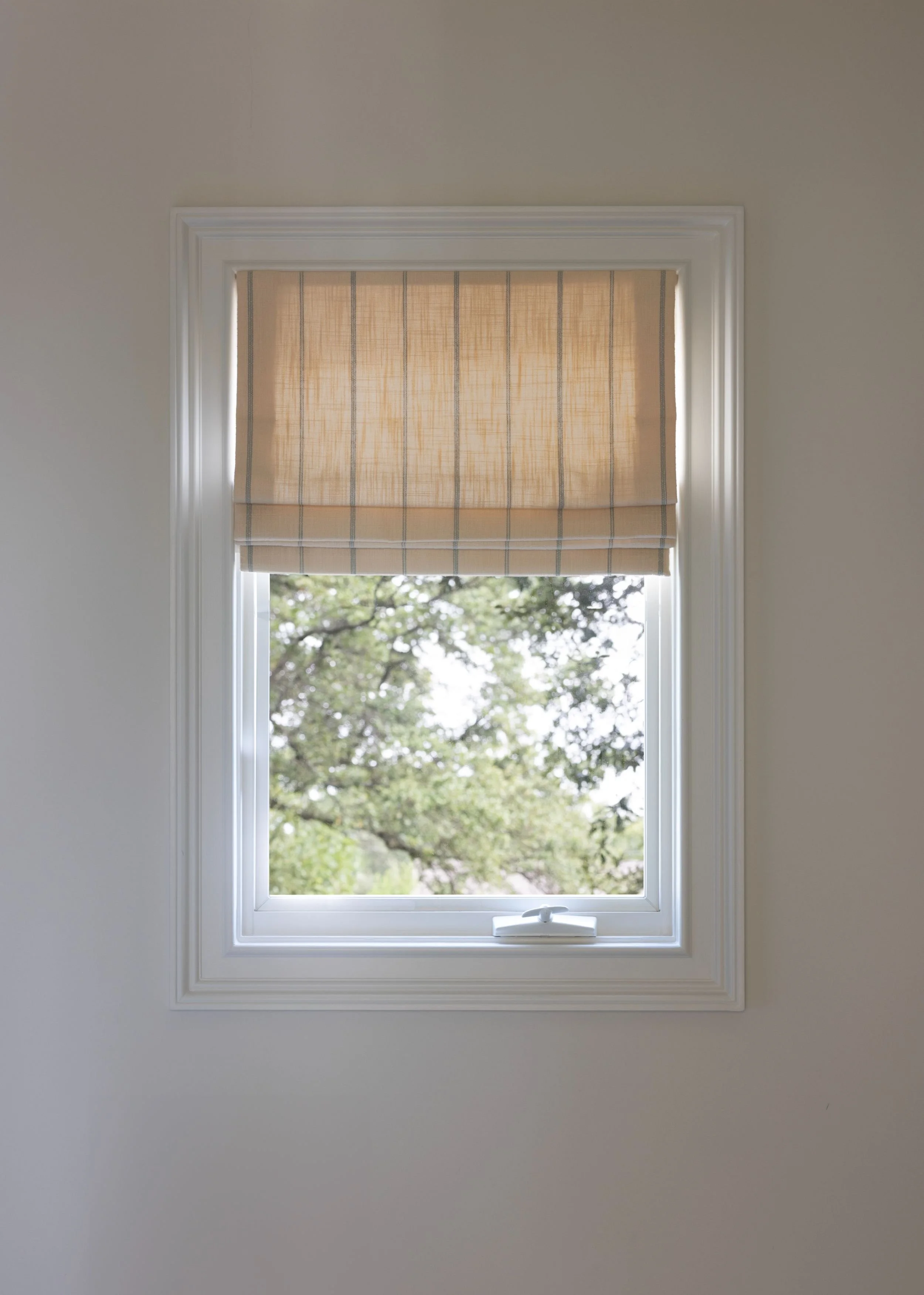 A window with a beige Roman shade and a view of green trees outside.