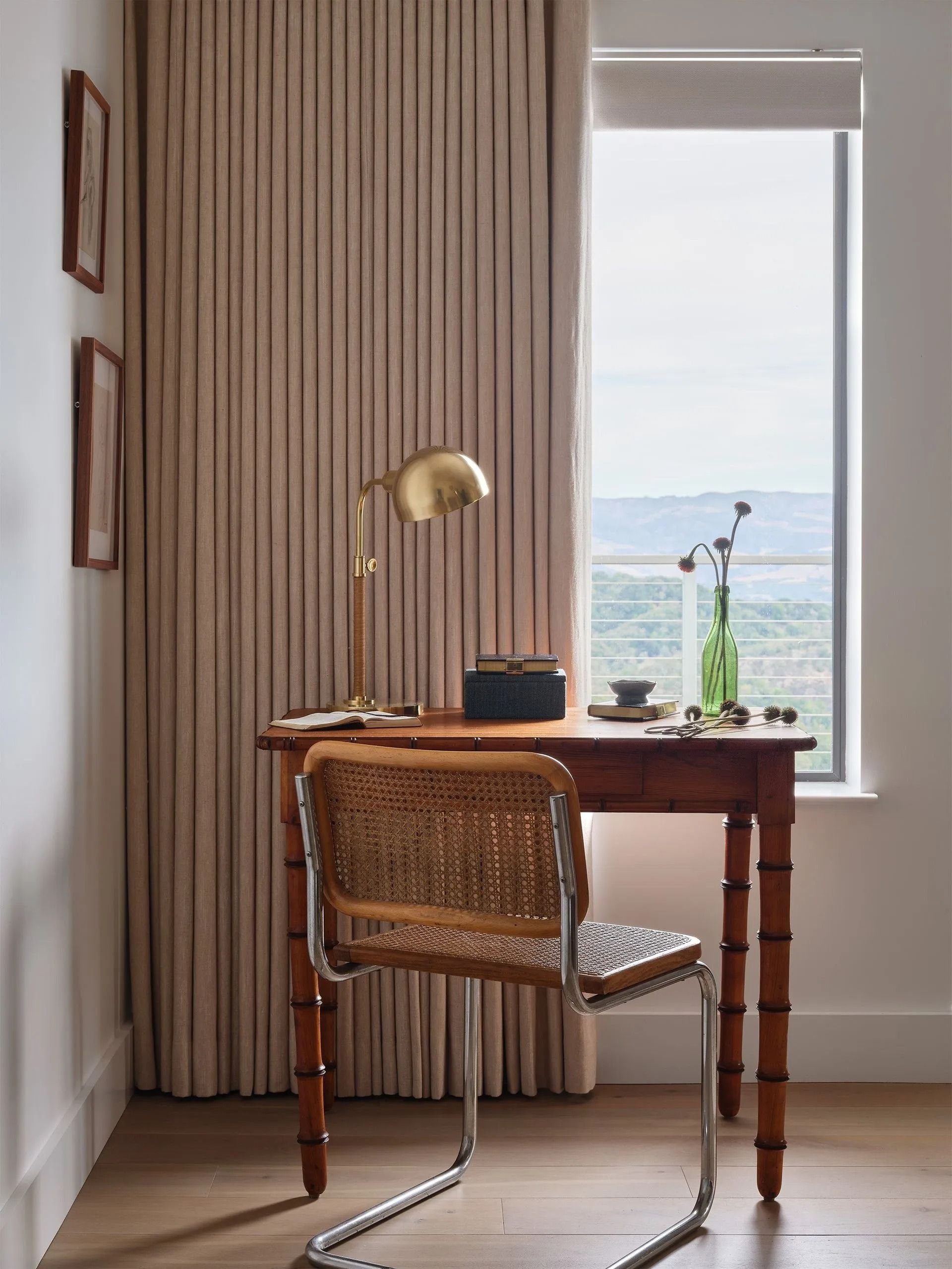 A cozy home office corner with a wooden desk, a vintage chair, a brass desk lamp, open book, black box, and a green vase with flowers, set next to a window with a view of distant hills.