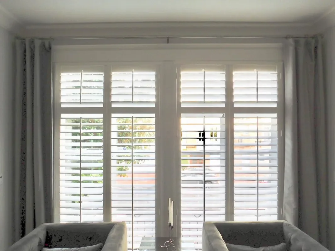 Bright room with large glass door with white plantation shutters, flanked by sheer white curtains, with two sofas partially visible at the bottom.