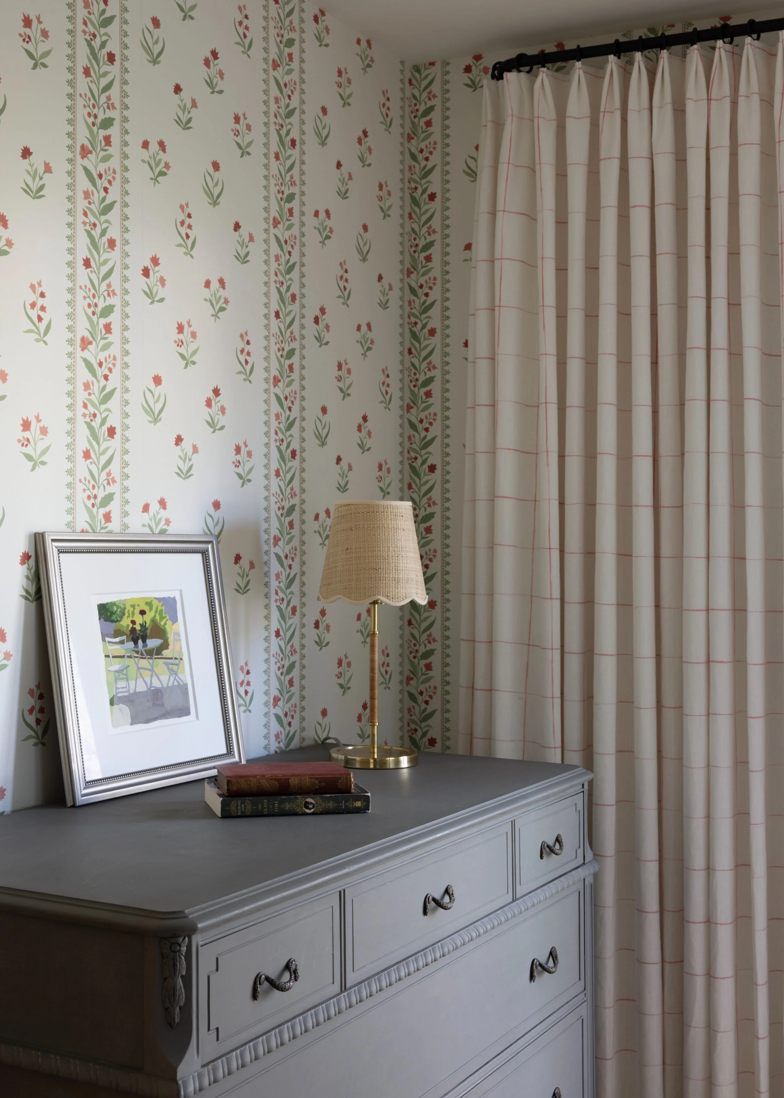 A white dresser with decorative trim, topped with a framed picture, a table lamp with a beige shade, and two books stacked on top. The background features floral wallpaper with red flowers and green leaves, and a cream-colored curtain with red lines 